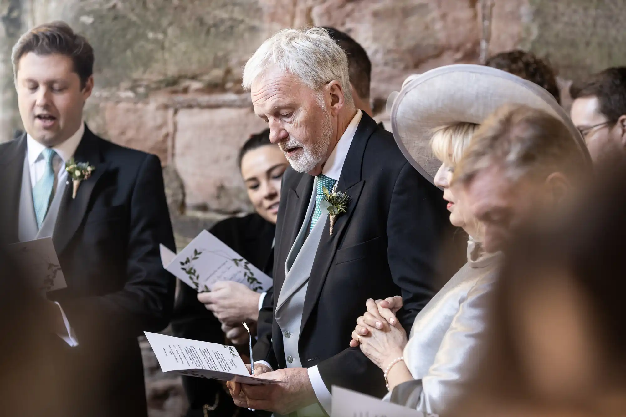 A group of people, some holding booklets, participate in a formal ceremony; one elderly man wears a suit with a boutonniere, and a woman beside him wears a grey outfit and hat.