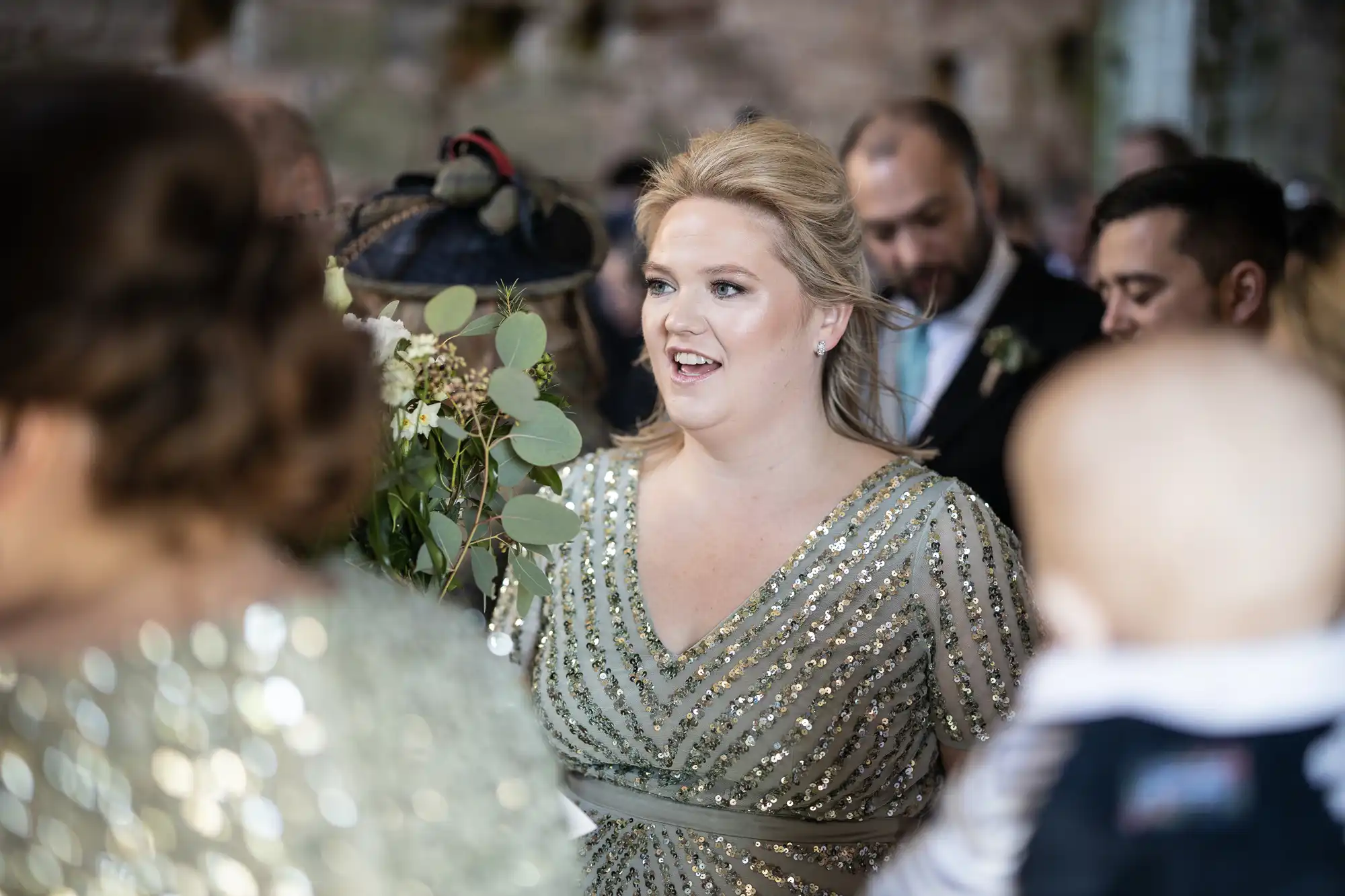 Woman in a sequined dress smiles while surrounded by people at an event. A man in a suit stands in the background.