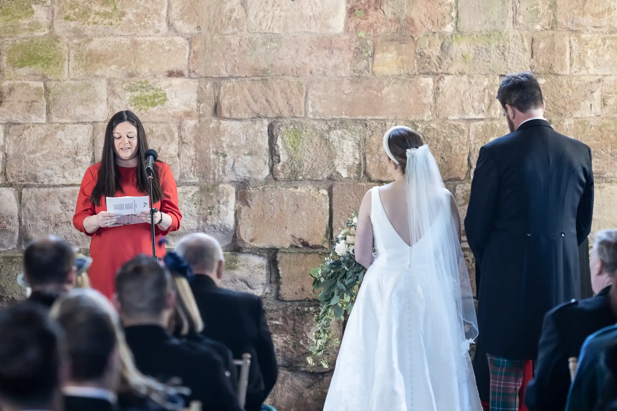 A woman in a red dress speaks at a microphone, while a bride in a white gown and a groom in a suit stand facing her against a rustic stone wall. Guests are seated in the foreground.