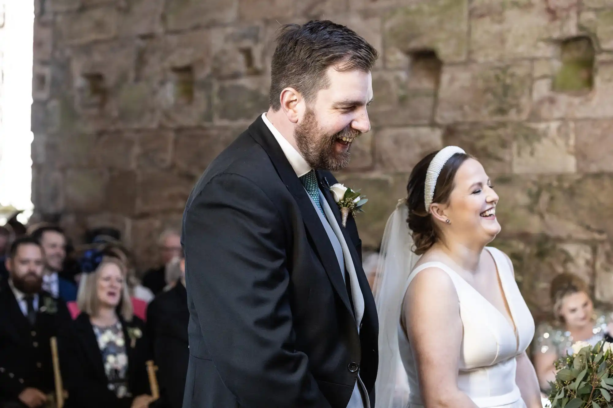 A bride and groom laugh during their wedding ceremony, surrounded by guests seated against a stone wall background.