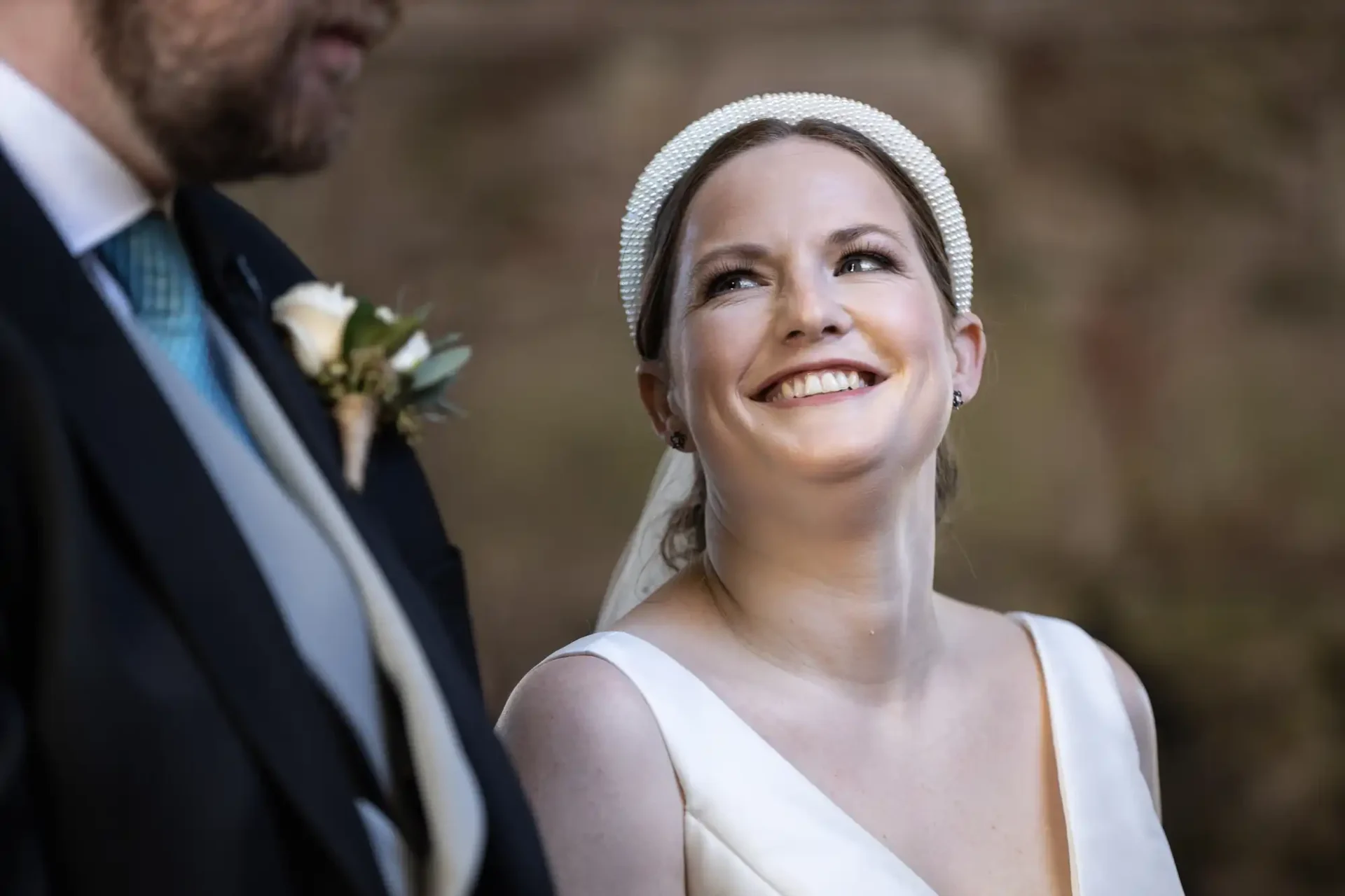 A bride in a white dress and headband smiles while looking at a groom in a suit and tie, adorned with a boutonniere.