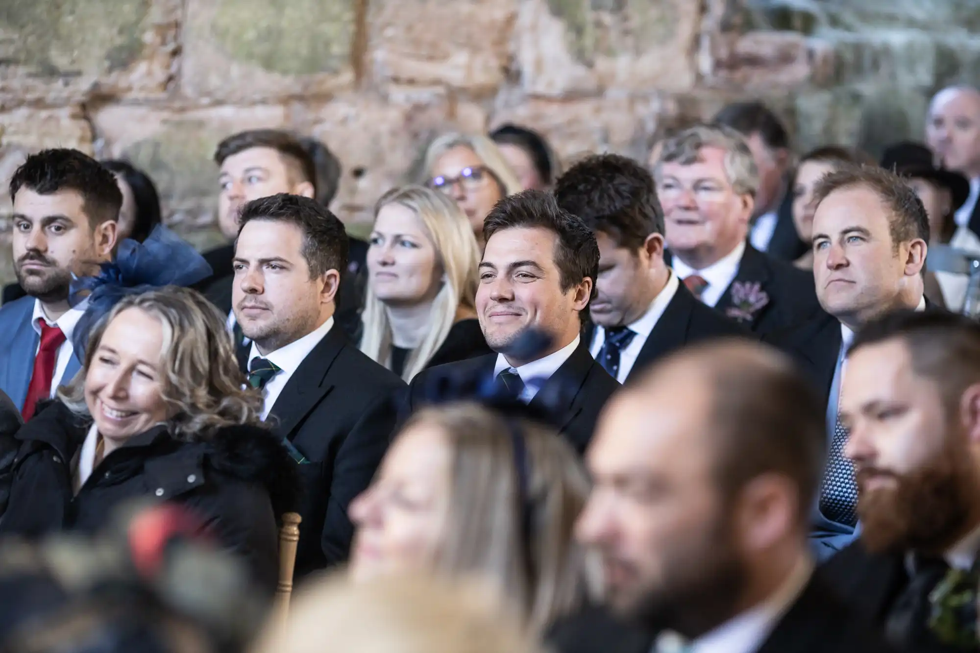 A group of people dressed formally, seated in rows, appear to be attending an event. Many are smiling while looking towards something out of the frame. The background features a stone wall.