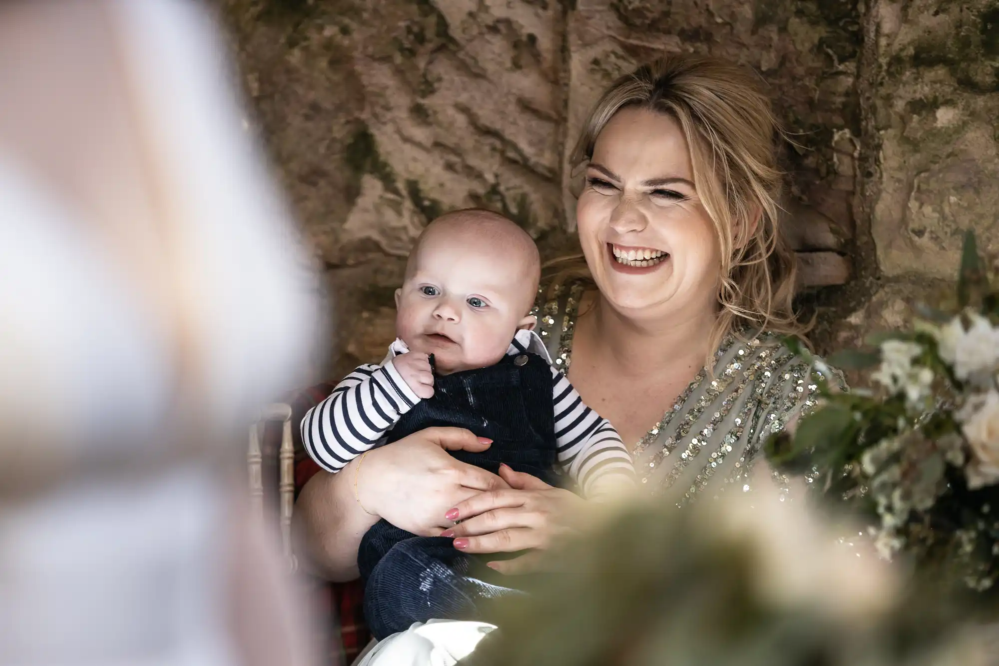 A woman is smiling and holding a baby. The baby is looking away, and the background shows a stone wall with some greenery.