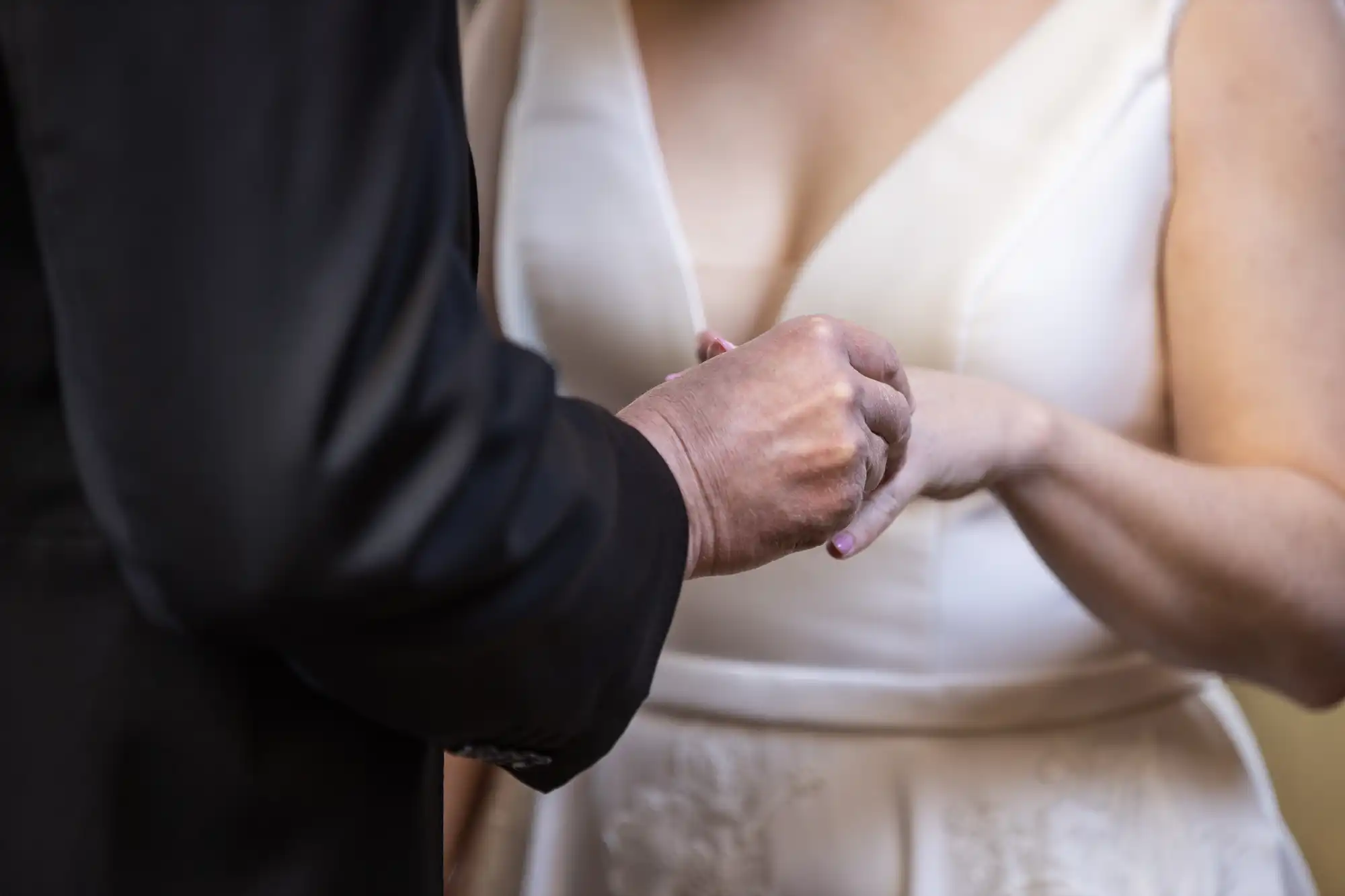 A close-up of two people holding hands, one in a black suit and the other in a white dress, likely during a wedding ceremony.