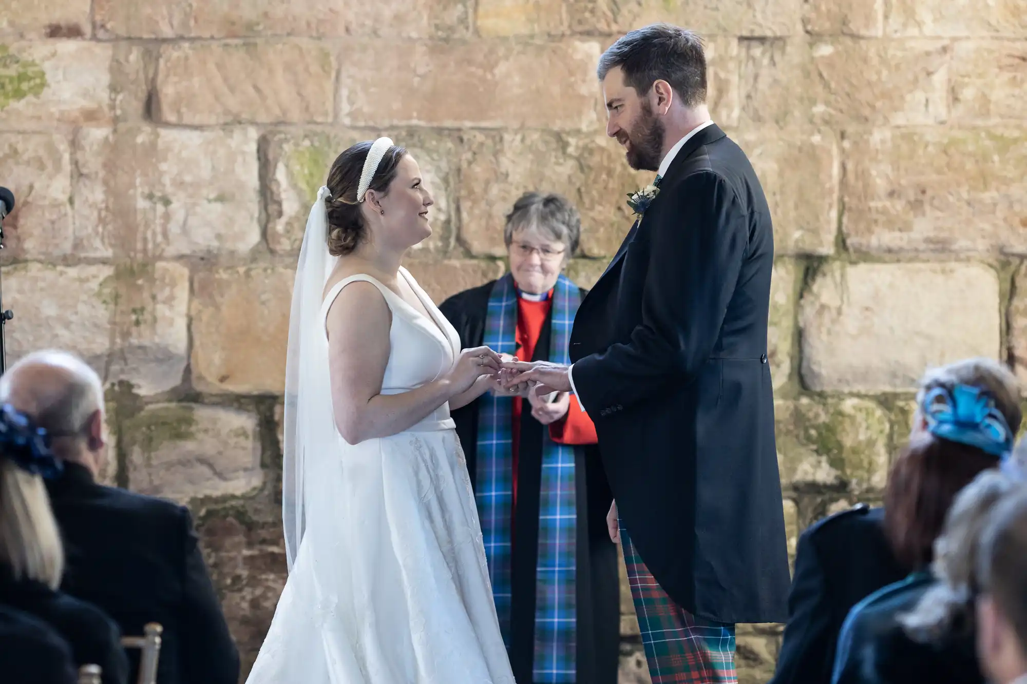 A couple is exchanging rings during a wedding ceremony. The bride is in a white dress, and the groom is in a suit with tartan pants. An officiant stands behind them against a stone wall backdrop.