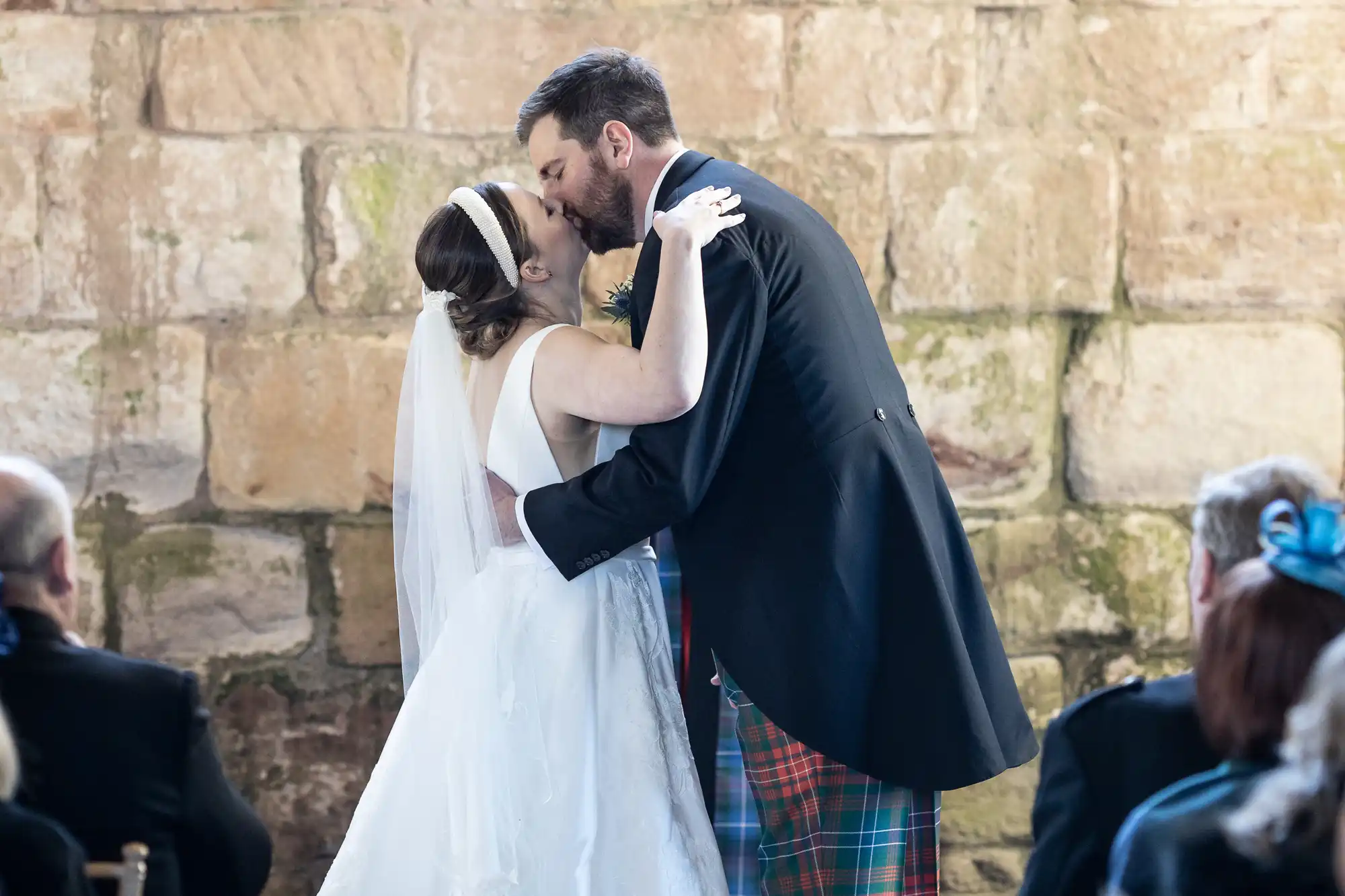 A bride and groom are kissing at their wedding ceremony in front of a stone wall. The bride is wearing a white dress and veil, and the groom is in a suit with a tartan kilt and a boutonniere.