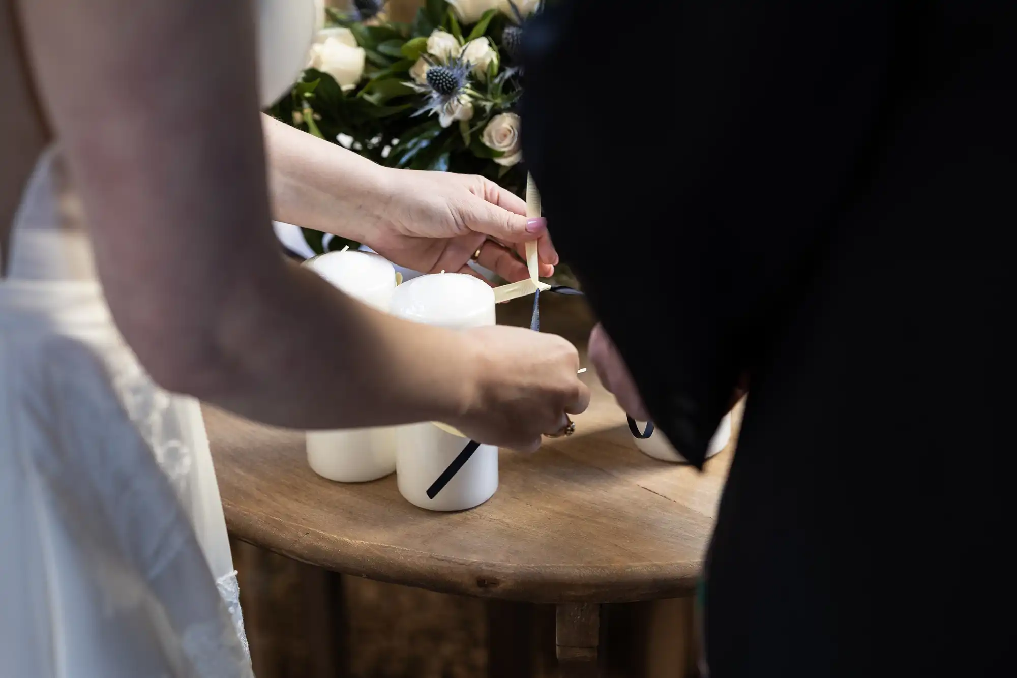 A person in formal attire lights a white candle on a wooden table, with another person partially visible beside them. Flowers are in the background.