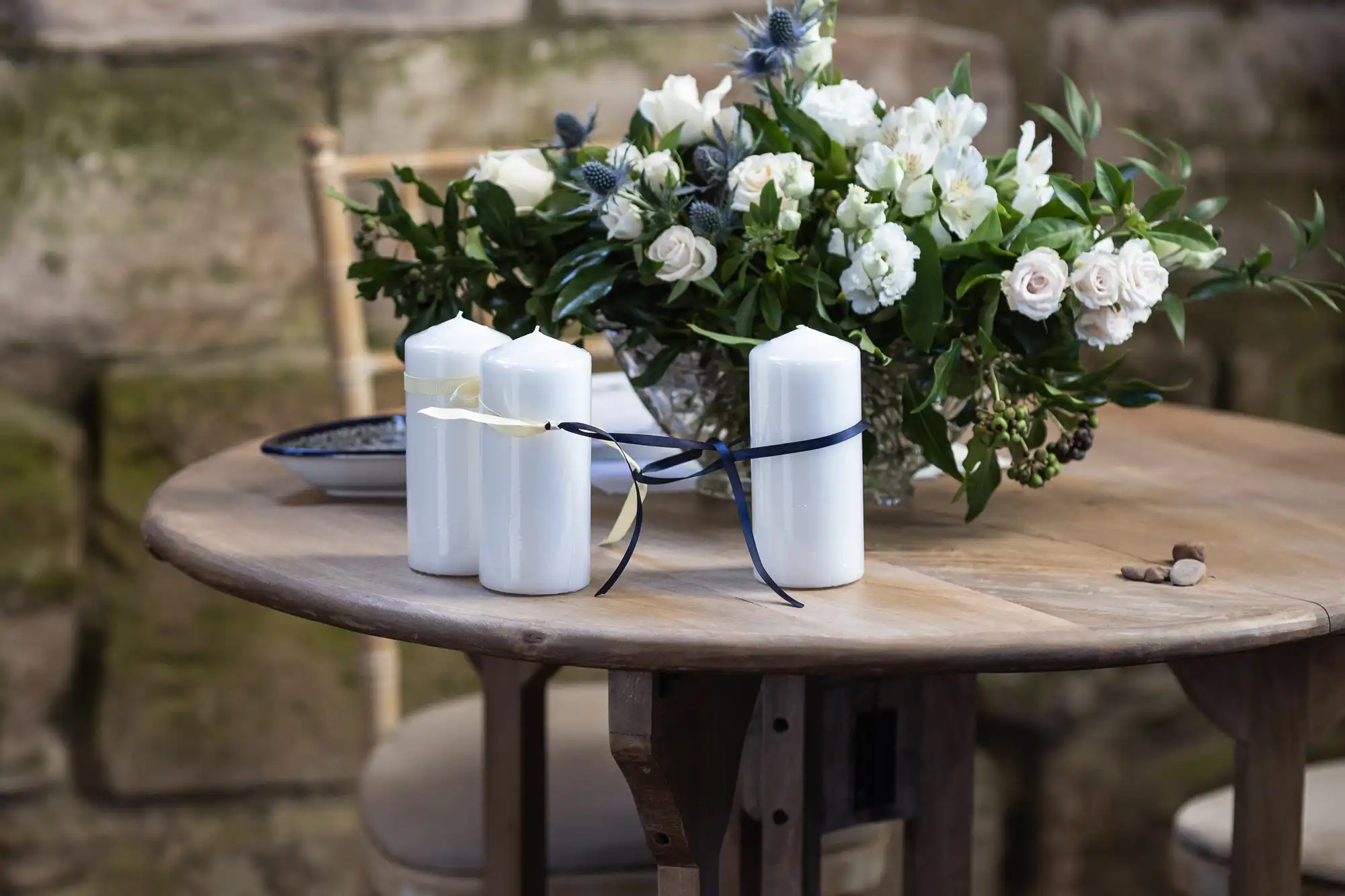 A wooden table holds four white candles with black and yellow ribbons and a floral centerpiece with white and green flowers.