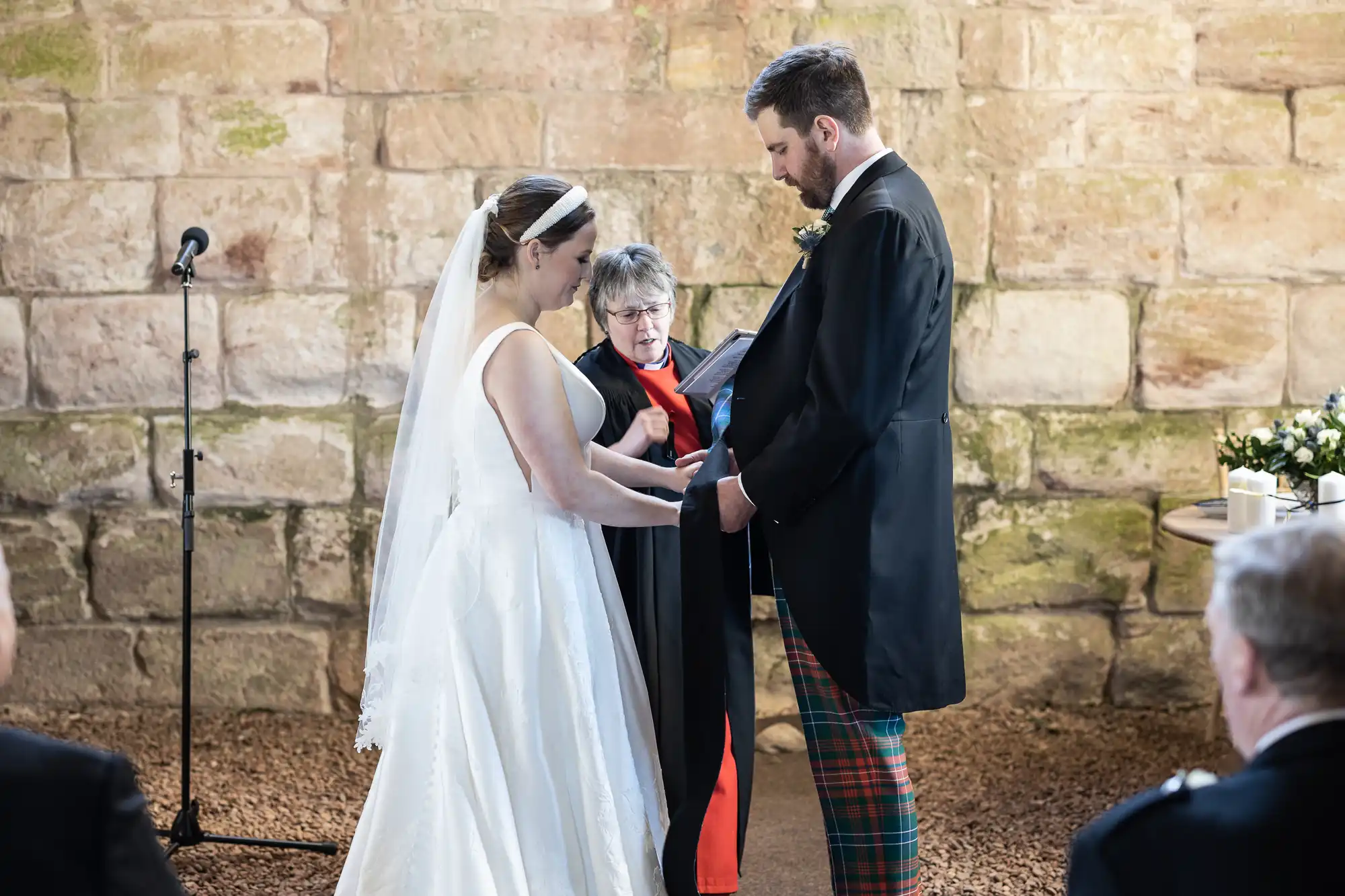 A bride and groom stand facing each other, holding hands, with a woman officiating their wedding ceremony in front of a stone wall. Guests are seated in the foreground.