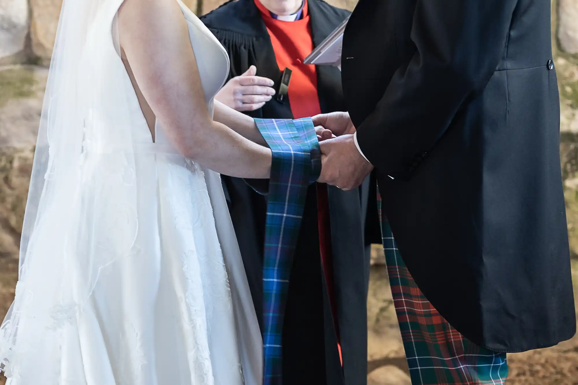 A couple standing at the altar, holding hands wrapped in a tartan cloth, during a wedding ceremony conducted by an officiant.