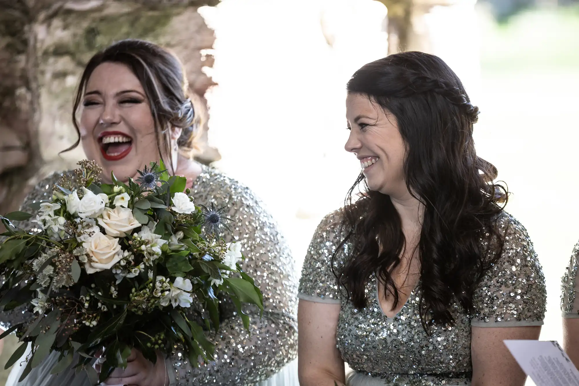 Two women wearing sequined dresses are captured smiling and laughing. One is holding a bouquet of flowers. The background appears to be an outdoor setting with natural light.