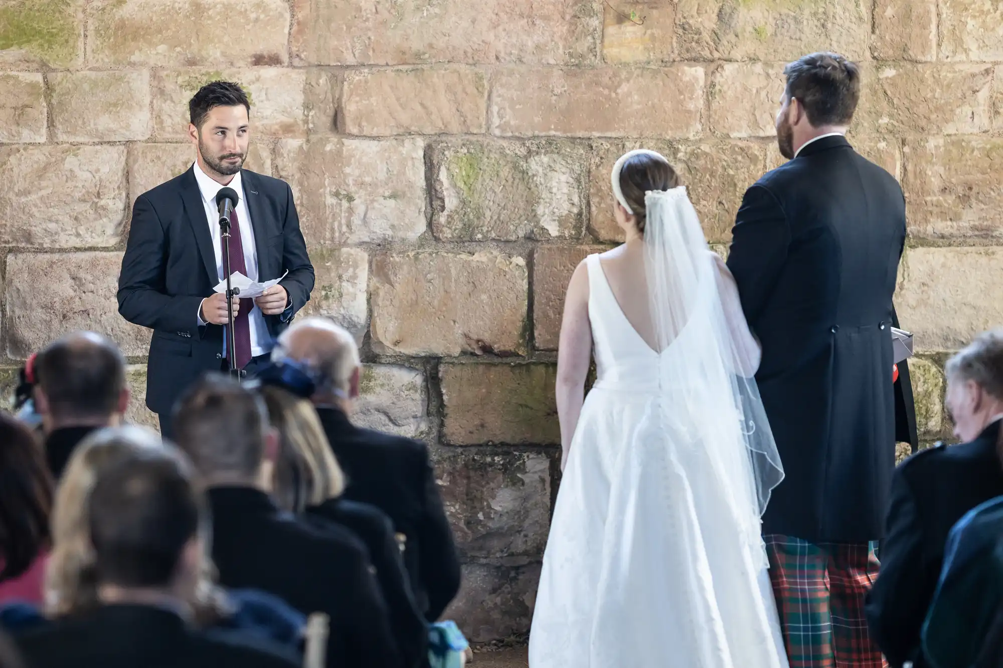 A man in a suit stands at a microphone holding notes, addressing a bride in a white dress with a veil and a groom in a dark suit with plaid pants, in front of a stone wall as guests watch.