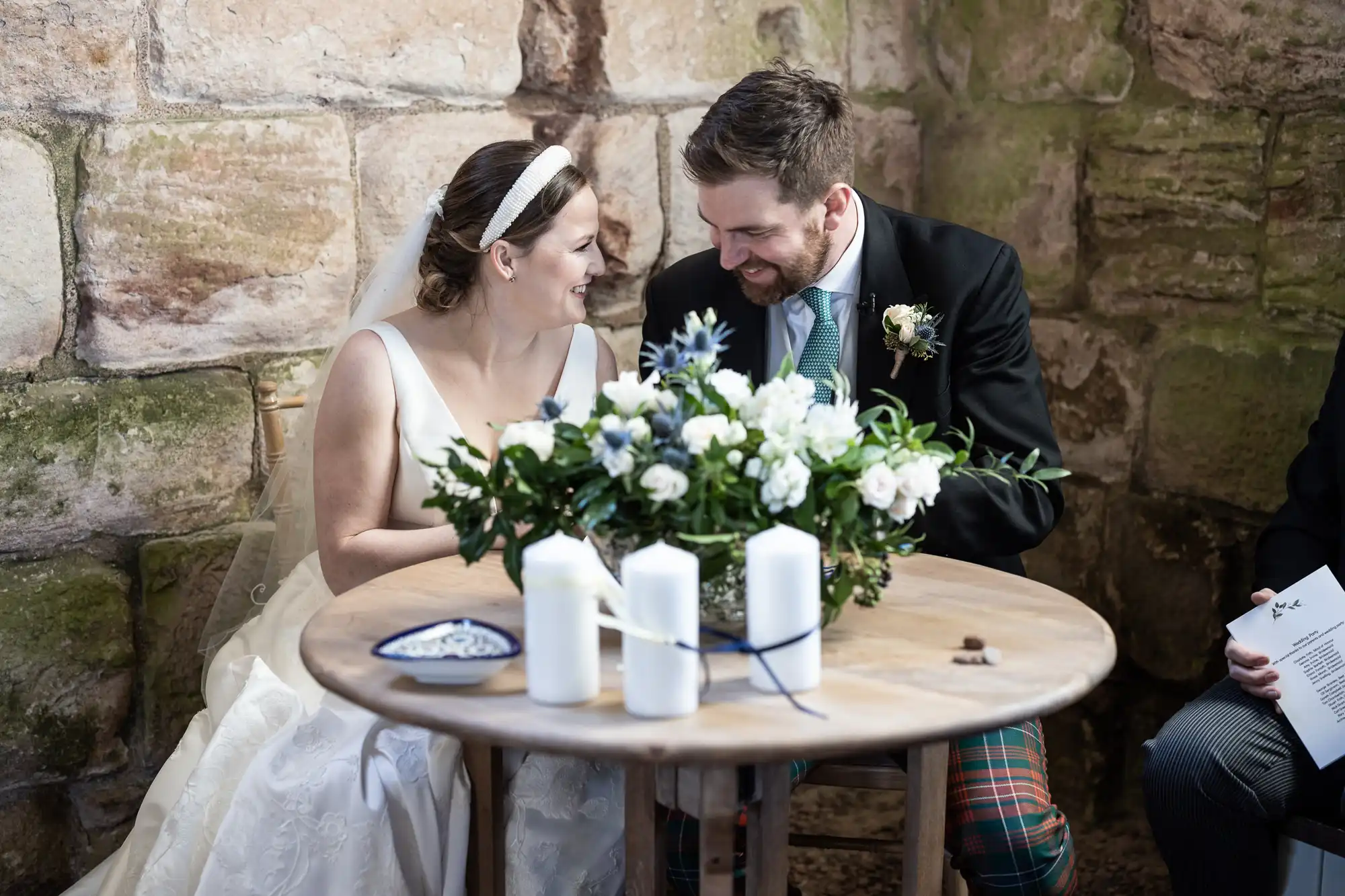 A bride and groom sit closely at a small wooden table adorned with white flowers and candles. They are smiling and leaning towards each other, sharing a joyful moment.