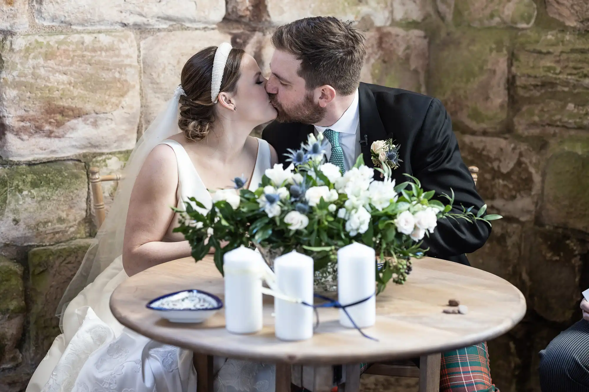 A bride and groom kiss while seated at a round table with white candles and floral arrangements.