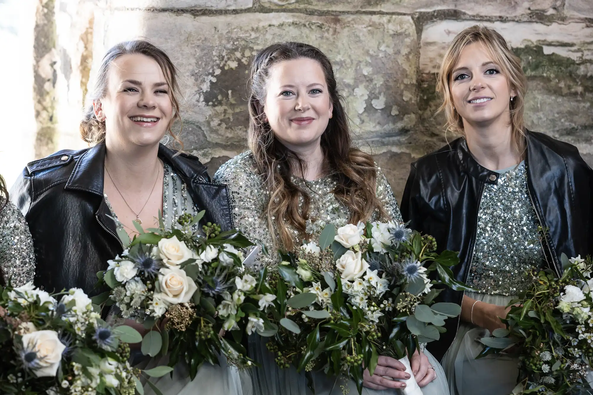 Three women in sparkling dresses and leather jackets hold flower bouquets, standing in front of a stone wall.