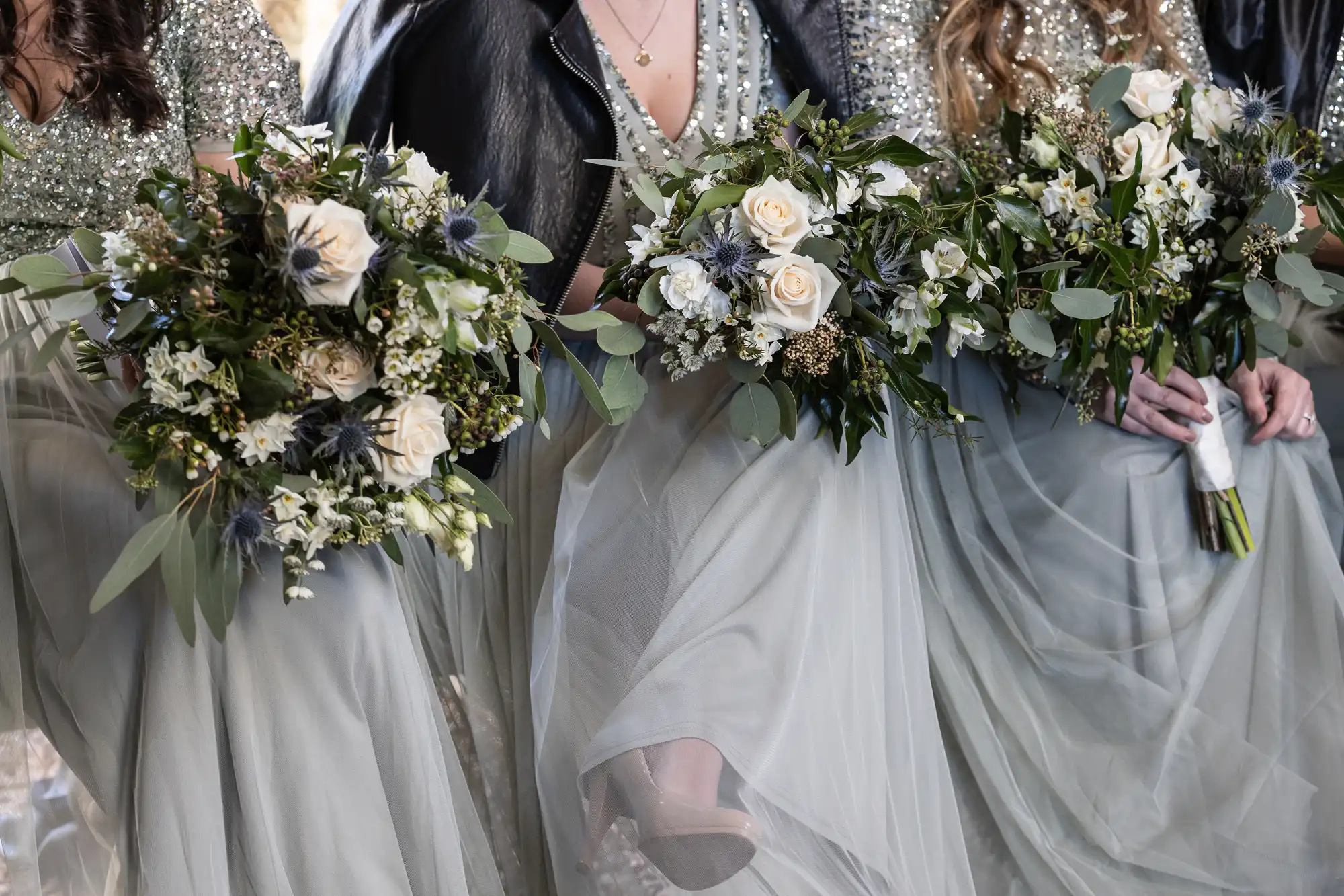 Close-up of bridesmaids holding floral bouquets with white and green flowers, wearing dresses with sheer skirts and glittery tops.