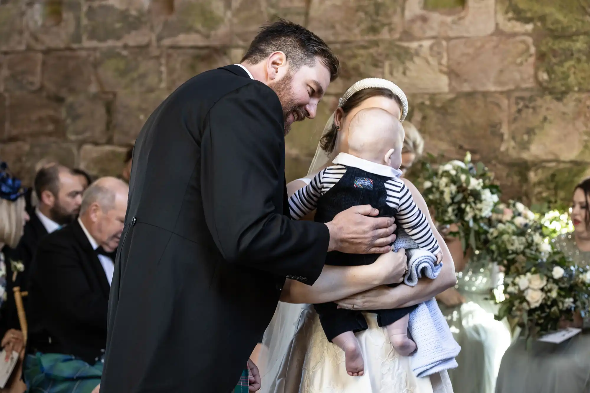 A bride holds a baby wrapped in a blue blanket while a man in a suit leans in to interact with the baby. Other people and a bouquet of flowers are visible in the background.