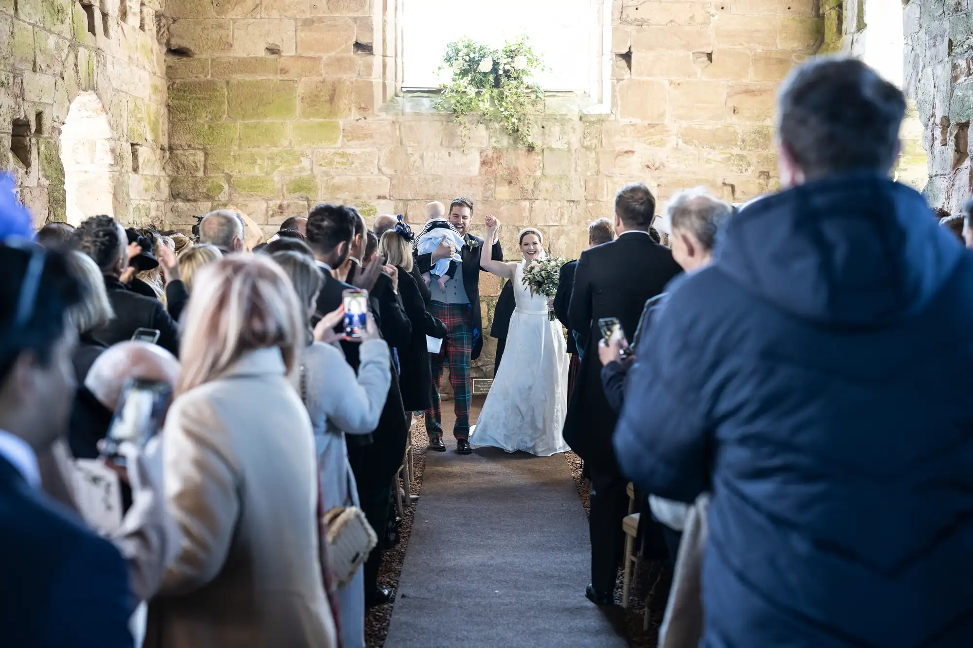 A bride and groom walk down the aisle at their wedding ceremony inside a rustic stone building, surrounded by guests taking photos.