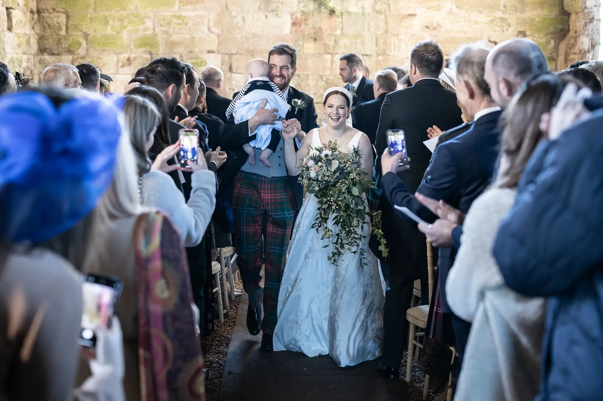 A newlywed couple walks down the aisle holding their baby, surrounded by guests taking photos and clapping in a stone-walled venue.