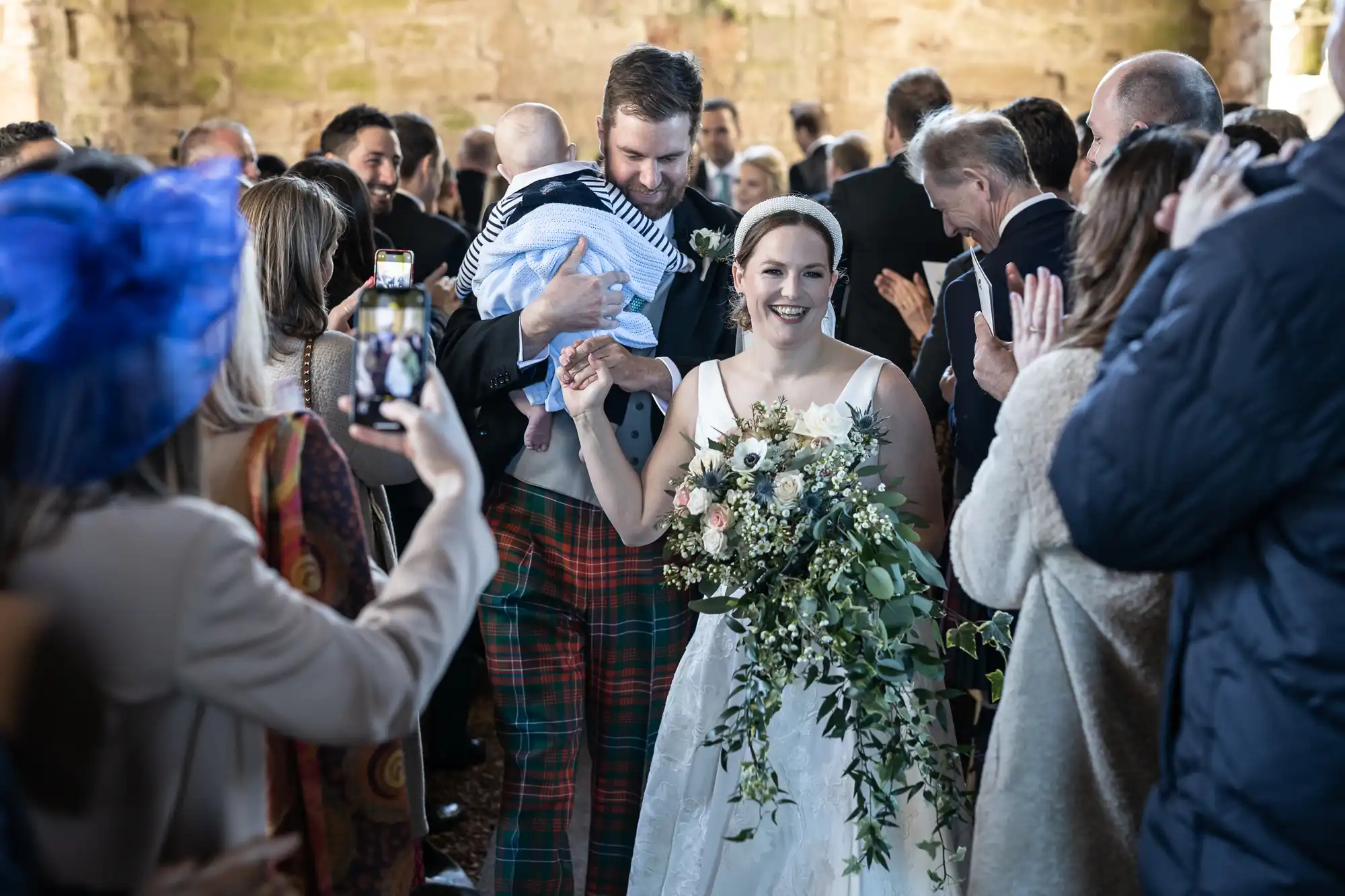 A bride and groom smile while leaving their wedding ceremony, surrounded by guests. The groom holds a baby, and some guests are taking pictures.