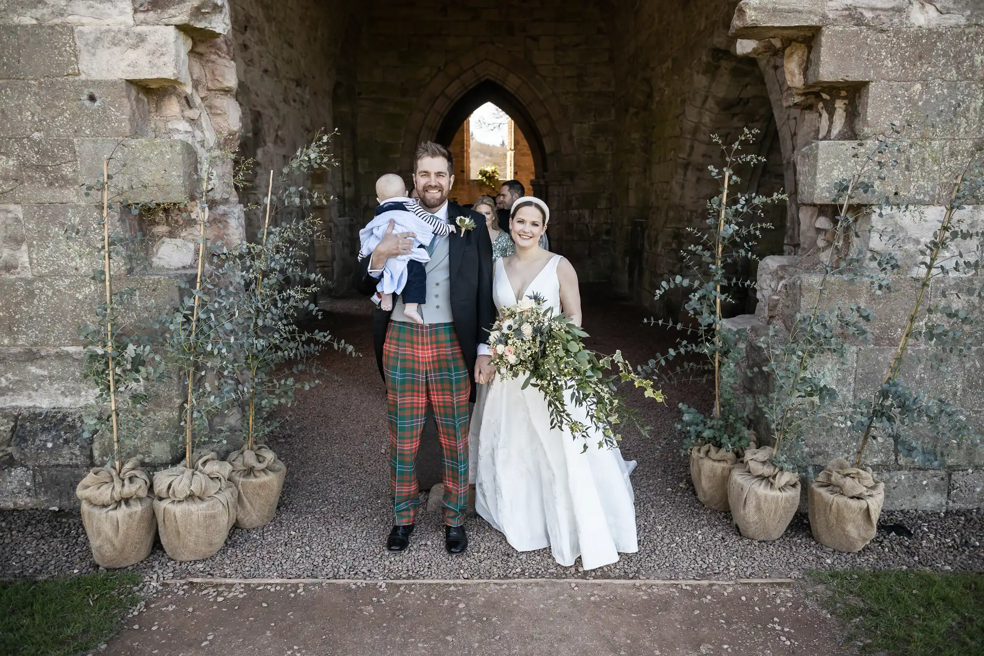 A couple in wedding attire stands at an archway, holding a baby. The man wears a kilt and jacket, while the woman wears a white gown and holds a bouquet.