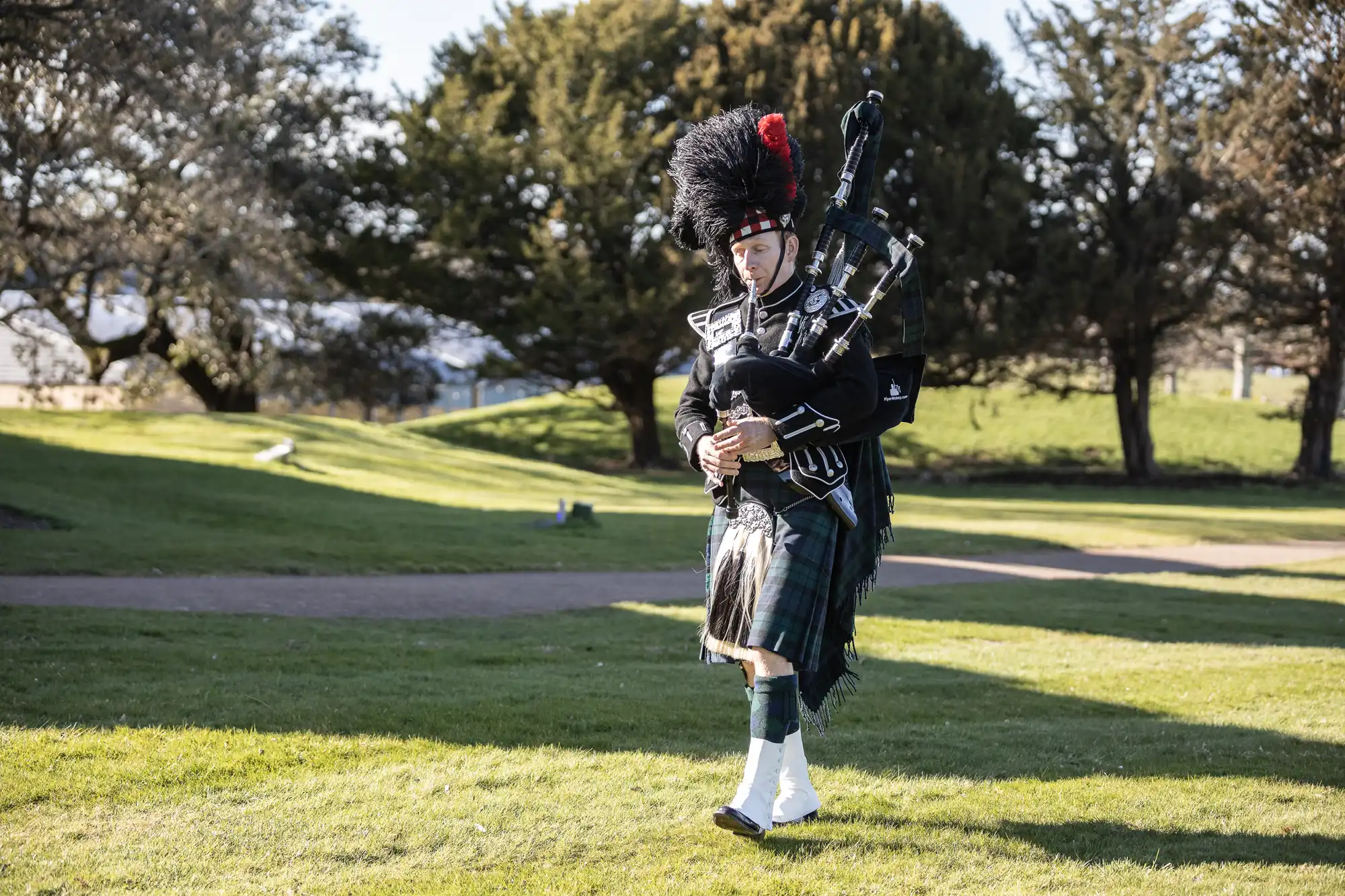 A person dressed in traditional Scottish attire plays the bagpipes while walking on a grassy field with trees in the background.