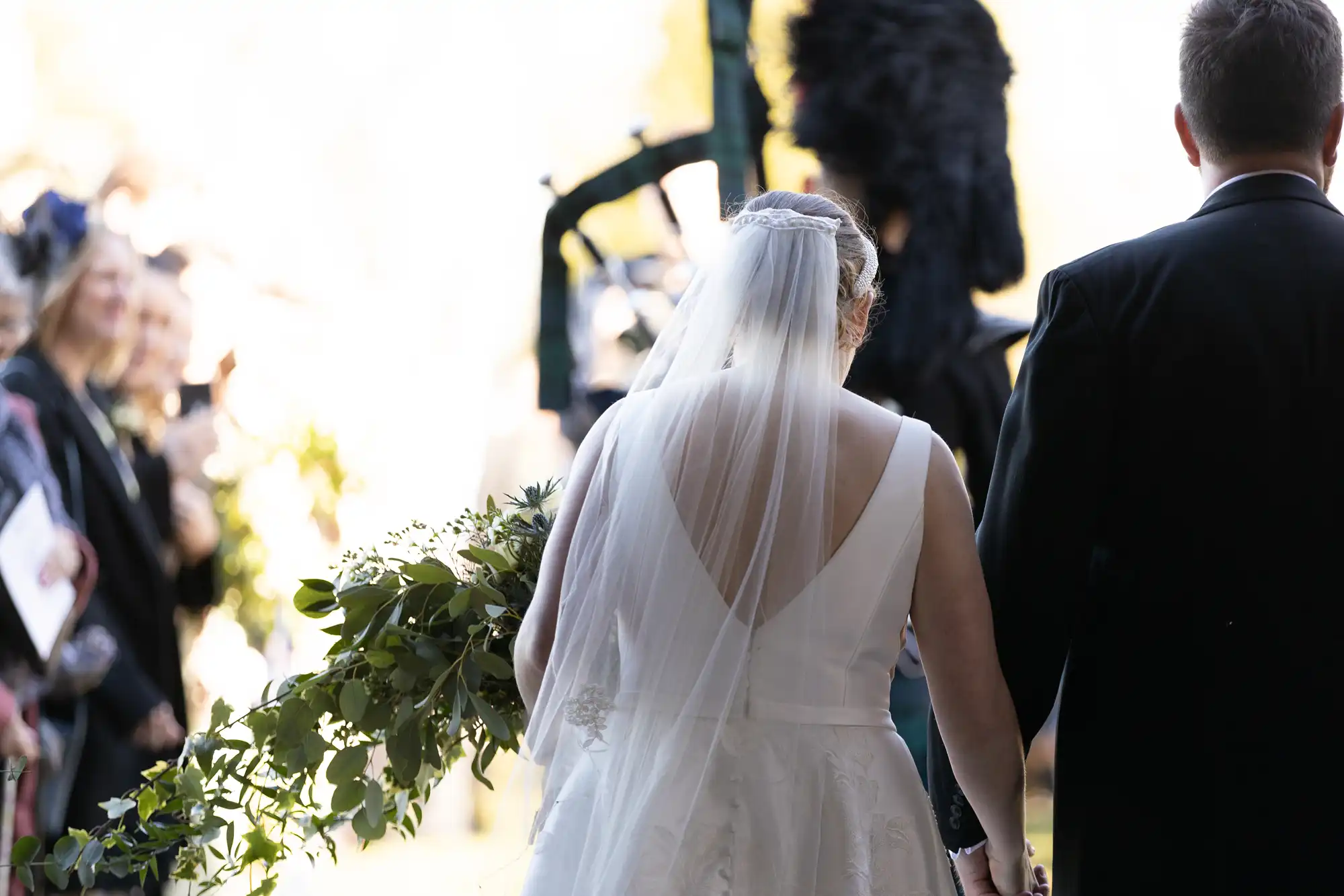 A bride and groom hold hands as they walk down the aisle during an outdoor wedding ceremony. The bride wears a white dress with a veil, and the groom is in a dark suit. Guests watch in the background.