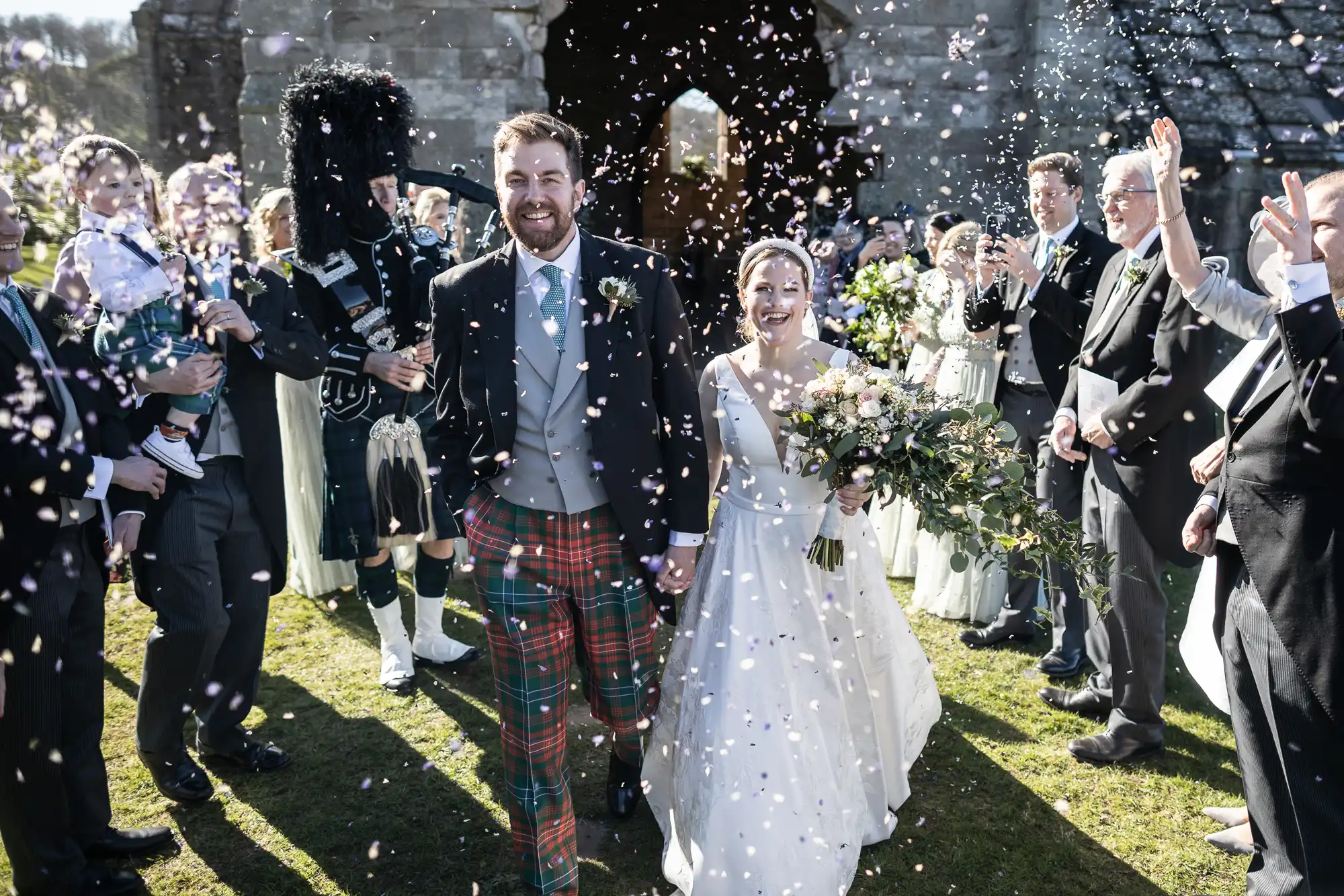 A bride and groom walk down the aisle outdoors, showered with confetti by guests. The groom is in a Scottish kilt, and a bagpiper is seen on the left.