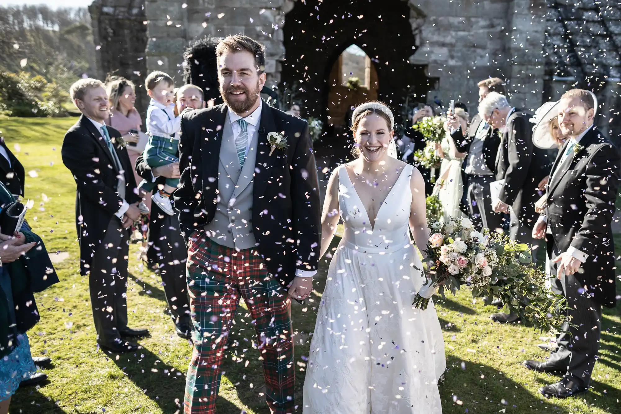 A newly married couple walks hand in hand as guests throw confetti. The groom wears a suit with plaid pants, while the bride wears a white dress and holds a bouquet of flowers.