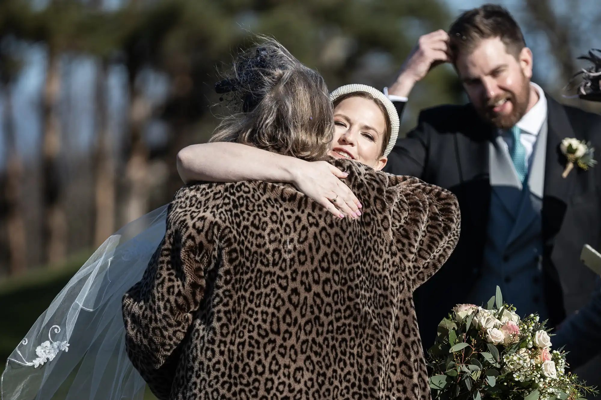 Bride in white dress hugs a person in a leopard print coat. A man in a suit stands nearby, adjusting his hair. They are outdoors, and the scene appears to be part of a wedding celebration.