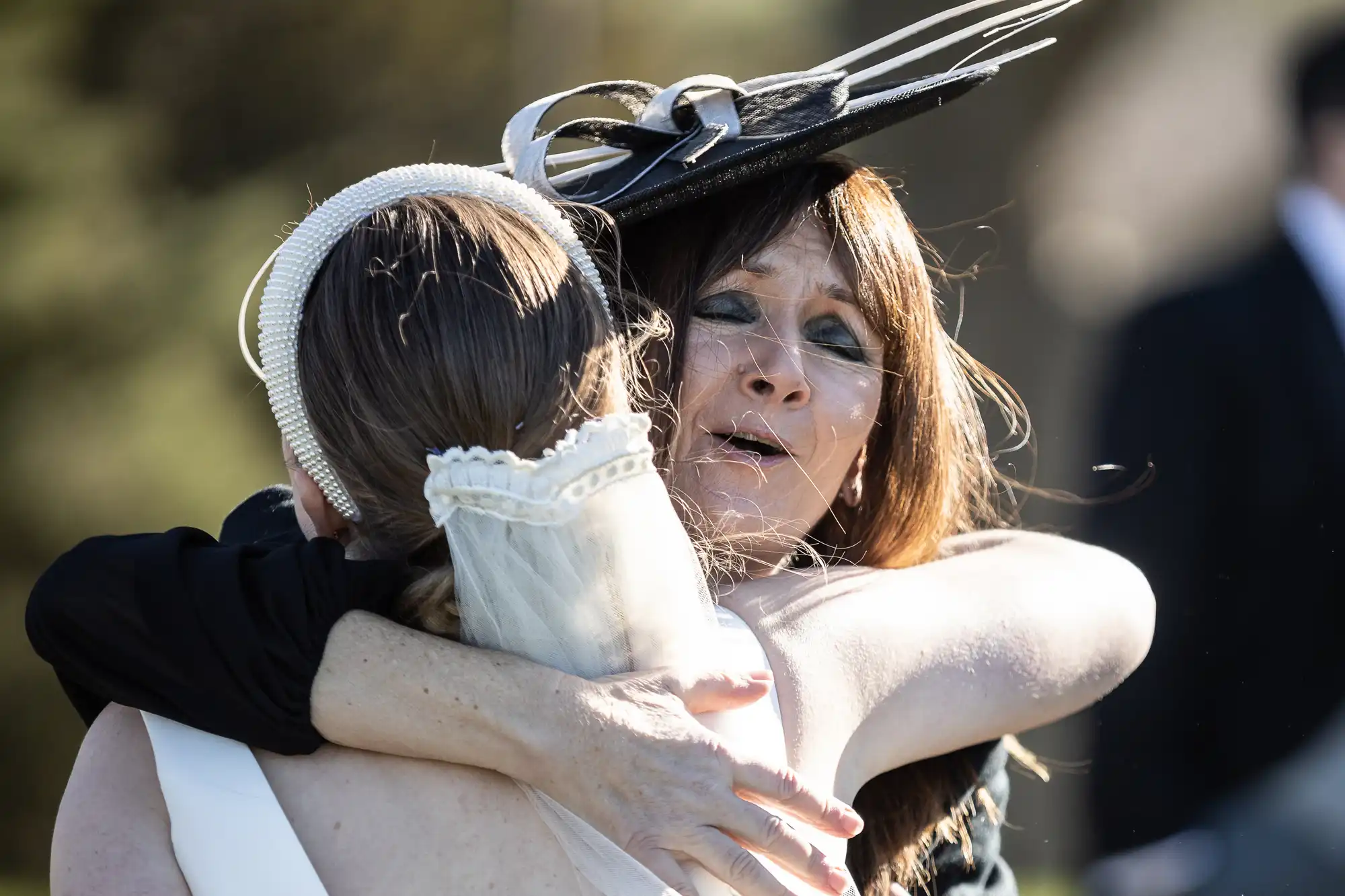 Two women are embracing, one in a wedding dress with a lace veil, the other in a black dress and a large black hat with decorative elements.