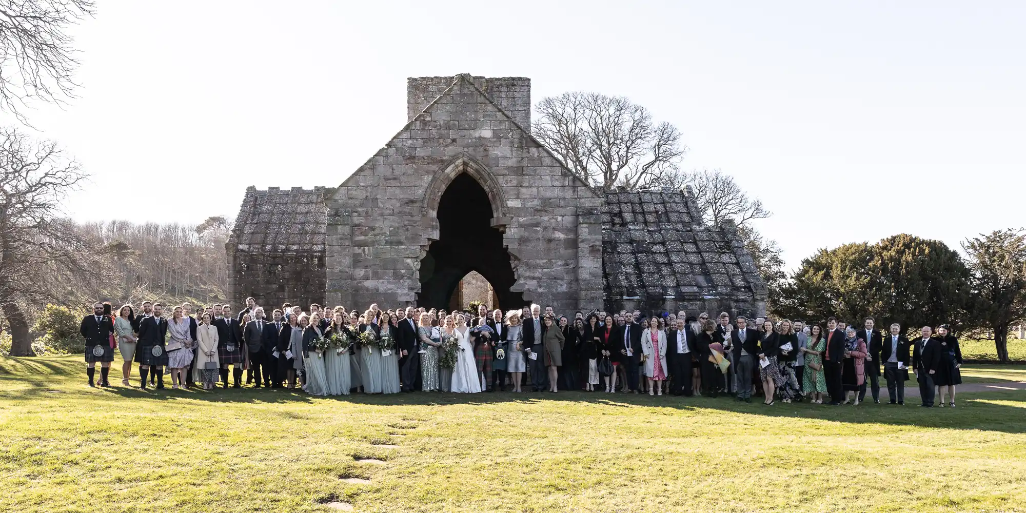 A large wedding party gathers for a group photo in front of a stone structure on a sunny day.