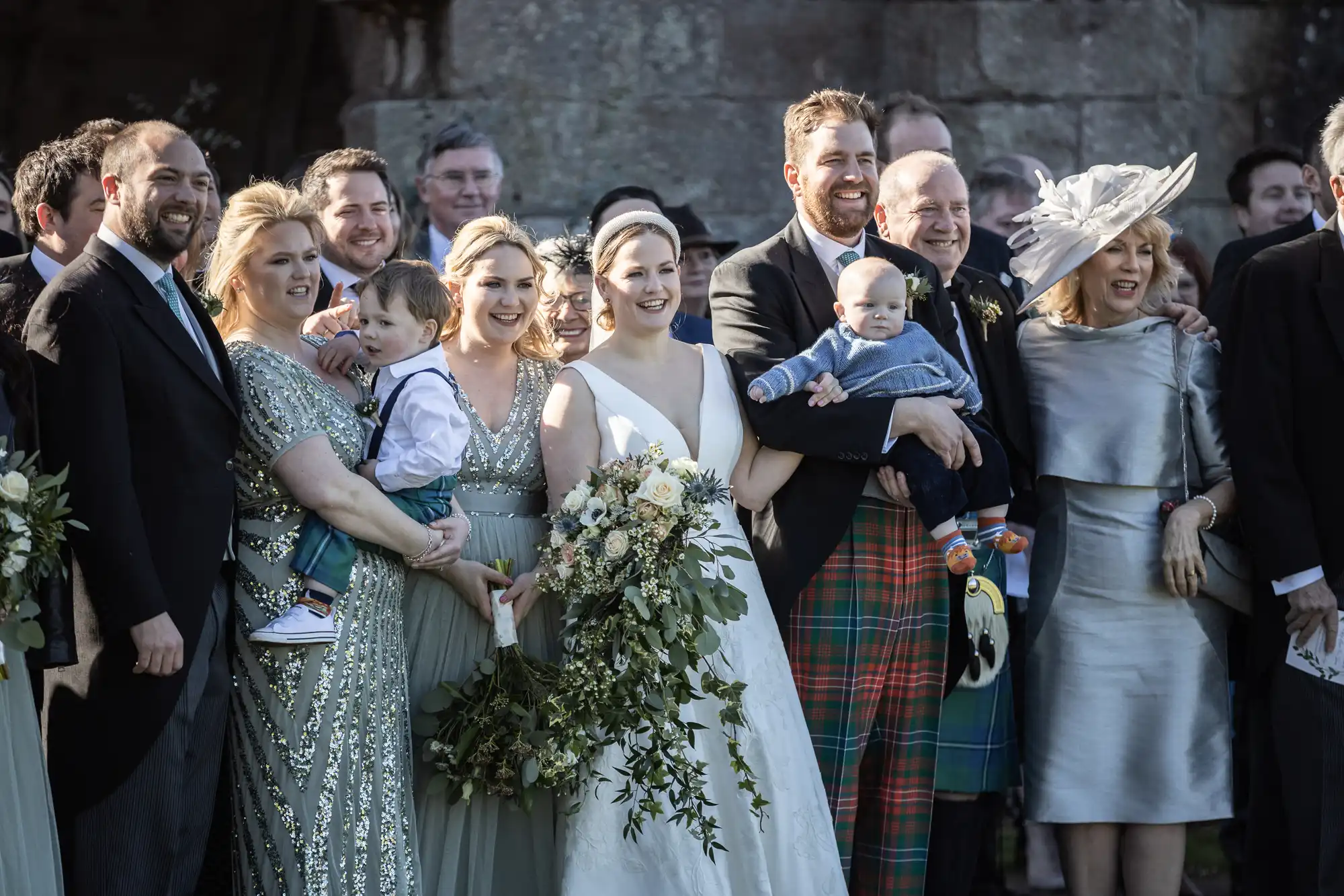 A wedding group portrait featuring the bride and groom with family and friends, standing closely together and smiling. The bride holds a bouquet, and two adults are holding toddlers.