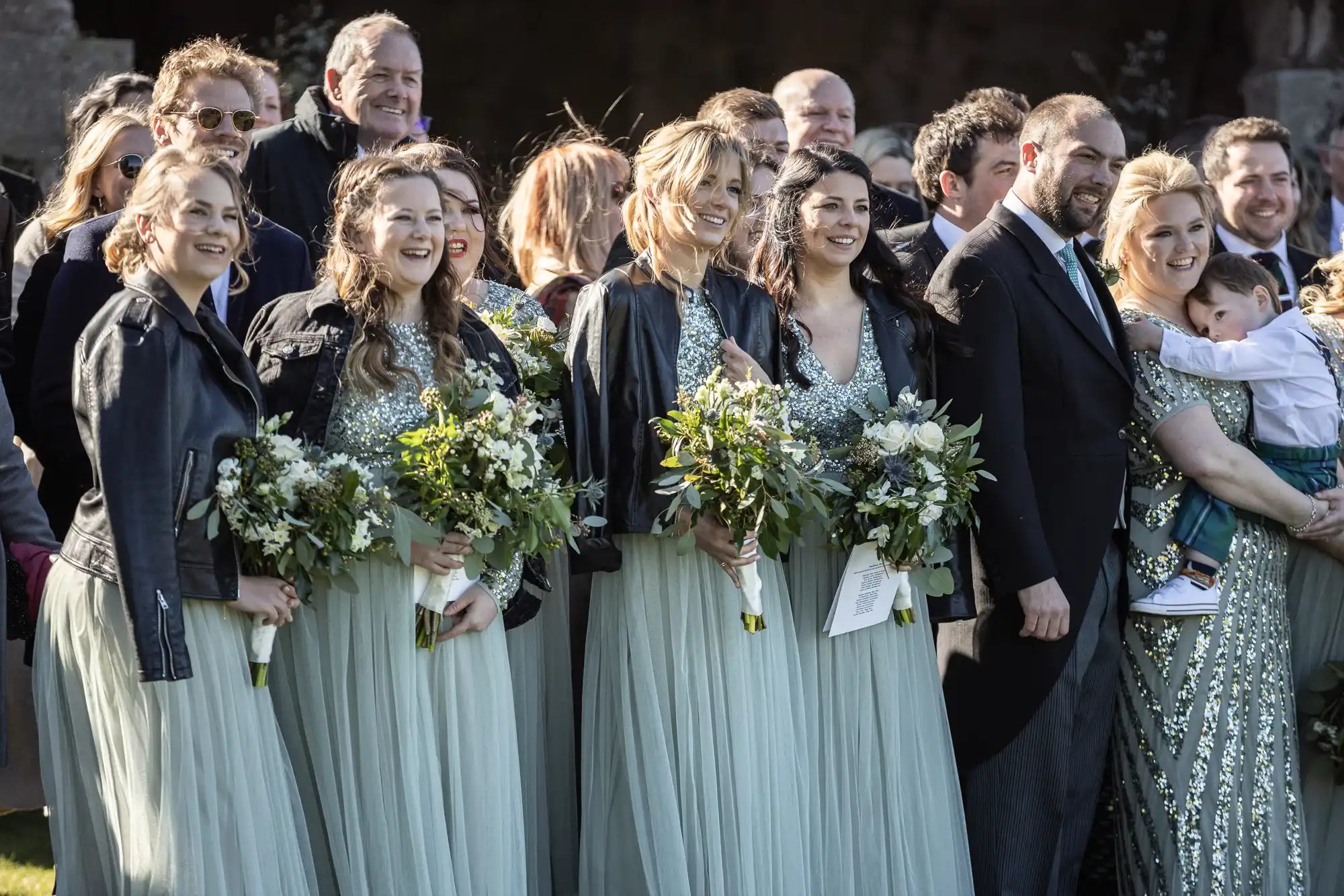 A group of bridesmaids in matching light green dresses and black jackets, holding bouquets, posing with other guests at an outdoor gathering.