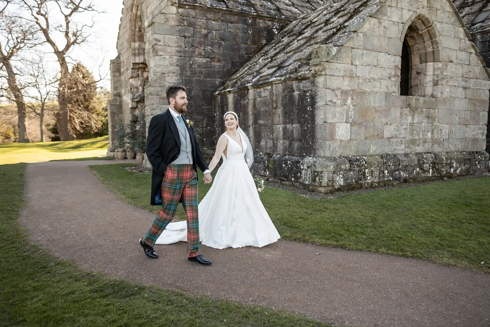 A bride in a white gown and a groom in a black suit with tartan pants walk hand in hand outside a stone building on a grassy path.