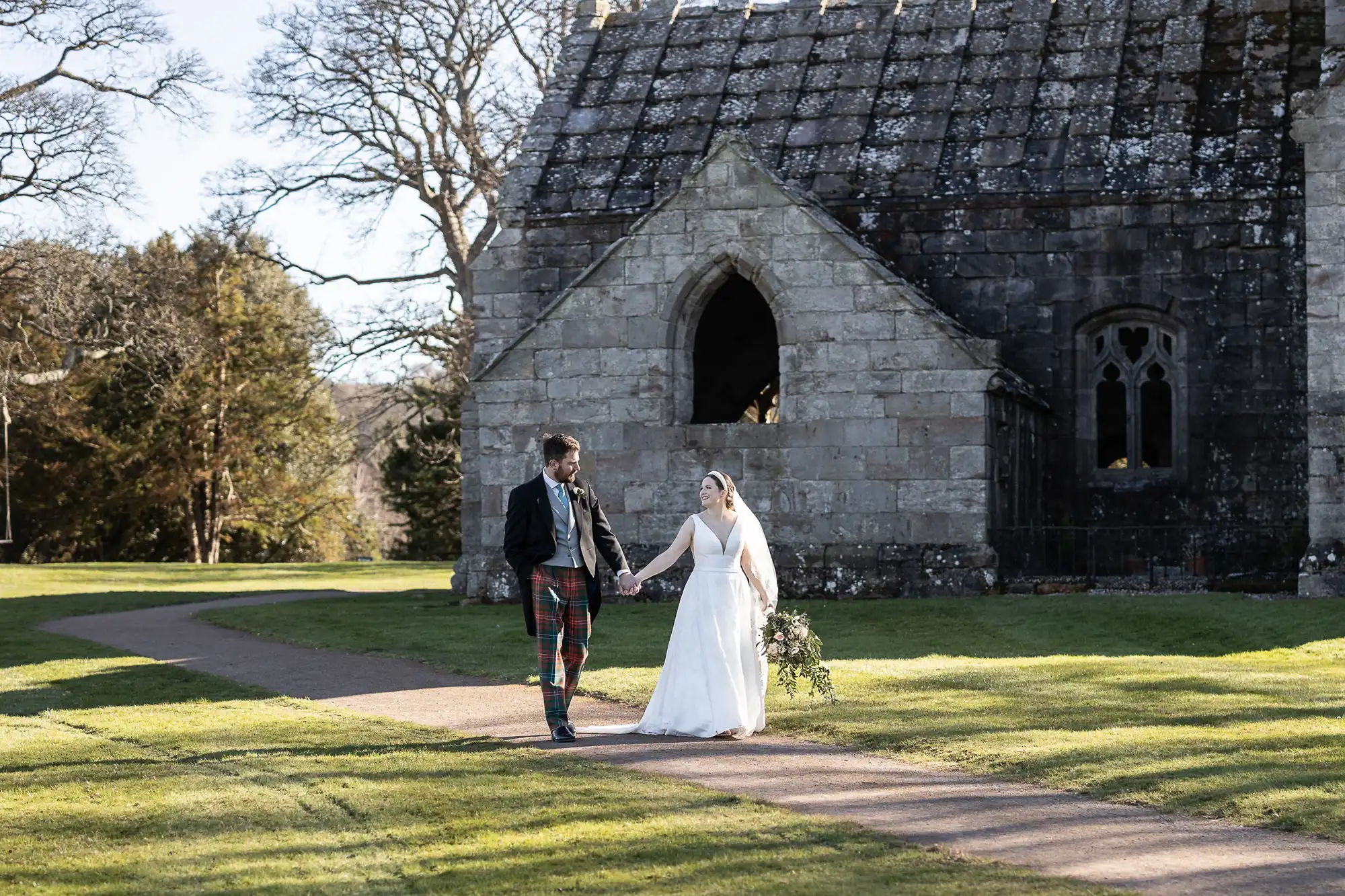 A couple, dressed in wedding attire, walks hand in hand on a path near an old stone church set in a grassy area with trees. The groom wears a kilt, and the bride holds a bouquet.