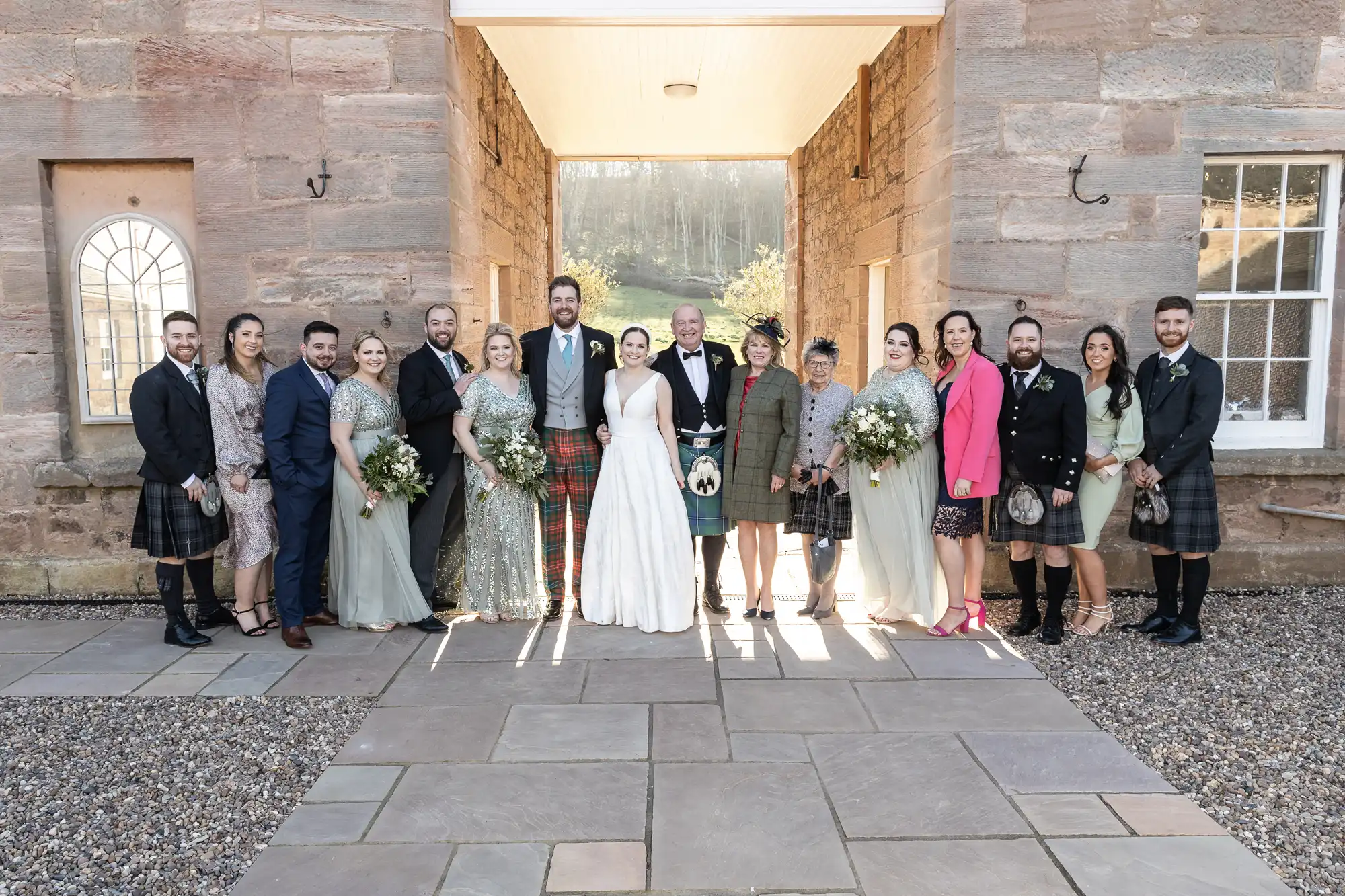 A wedding party stands together for a group photo. The bride and groom are centered, with bridesmaids and groomsmen on either side. Some are dressed in kilts, and the setting is outdoors in front of a stone building.