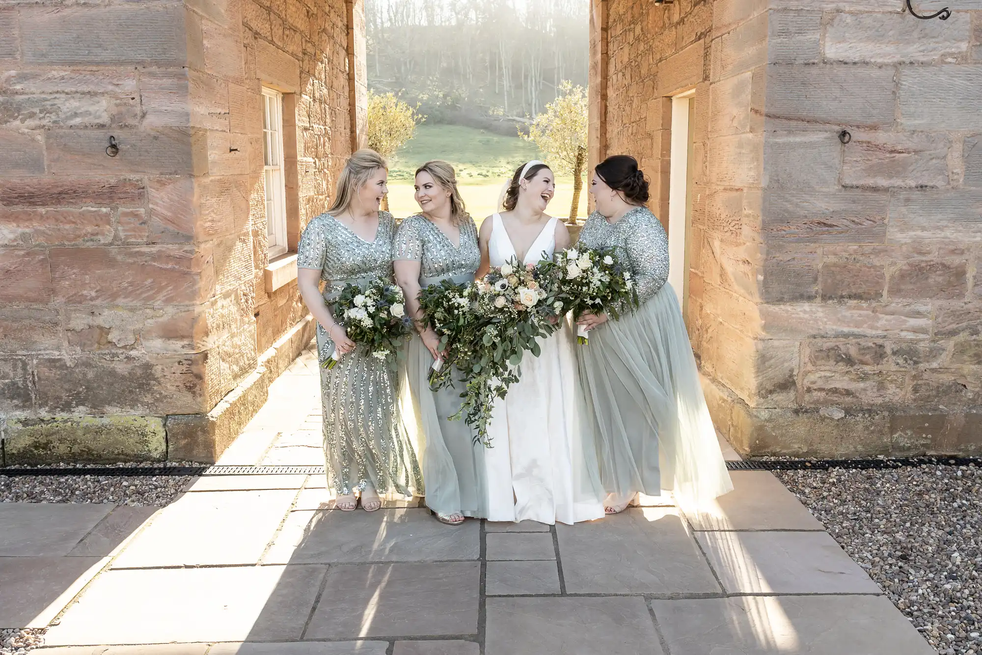 A bride in a white dress stands with three bridesmaids in matching silver-green dresses, holding large floral bouquets, between stone pillars on a sunny day.