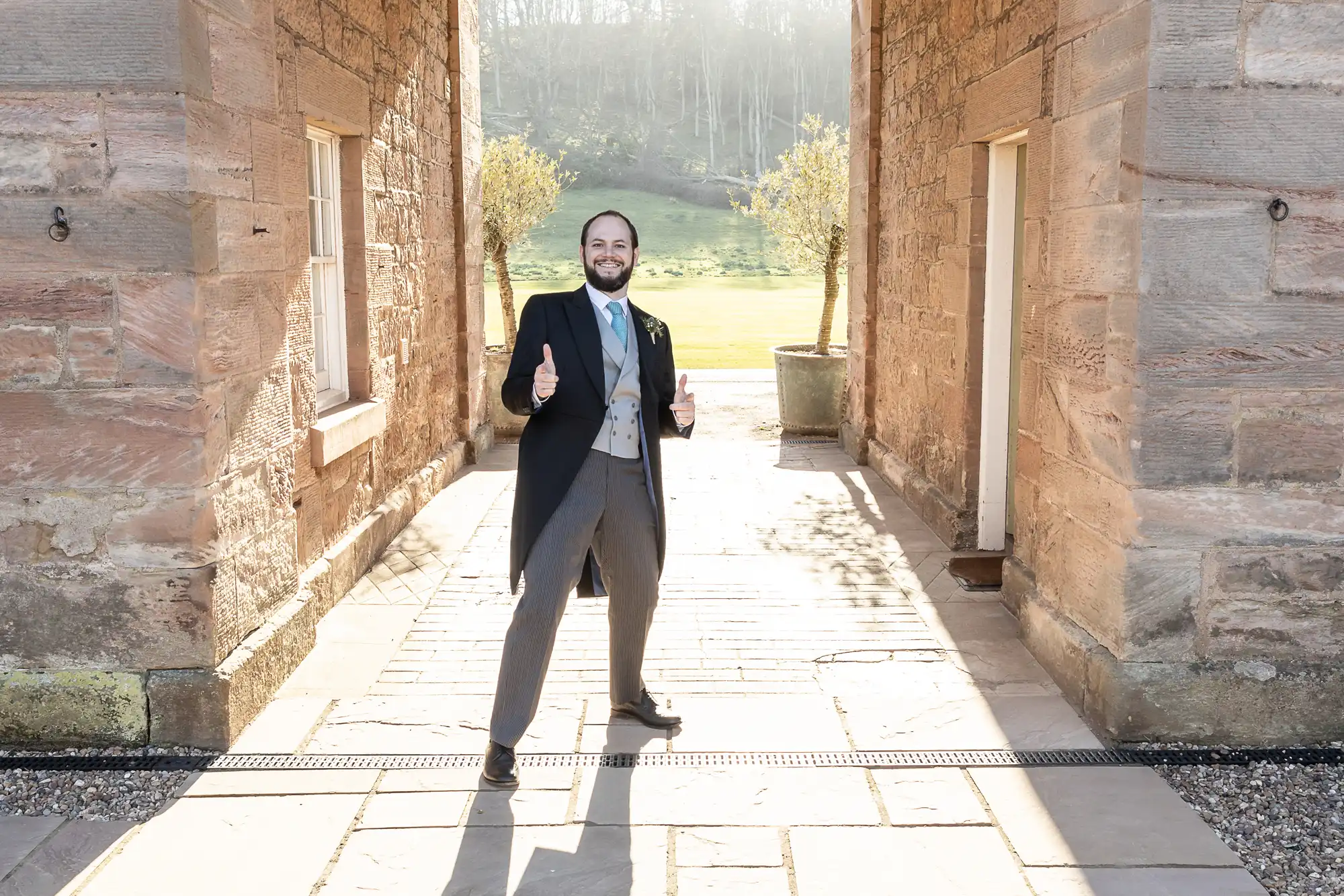 A man in formal attire stands outdoors between two stone walls, smiling and giving two thumbs up in a sunlit area.