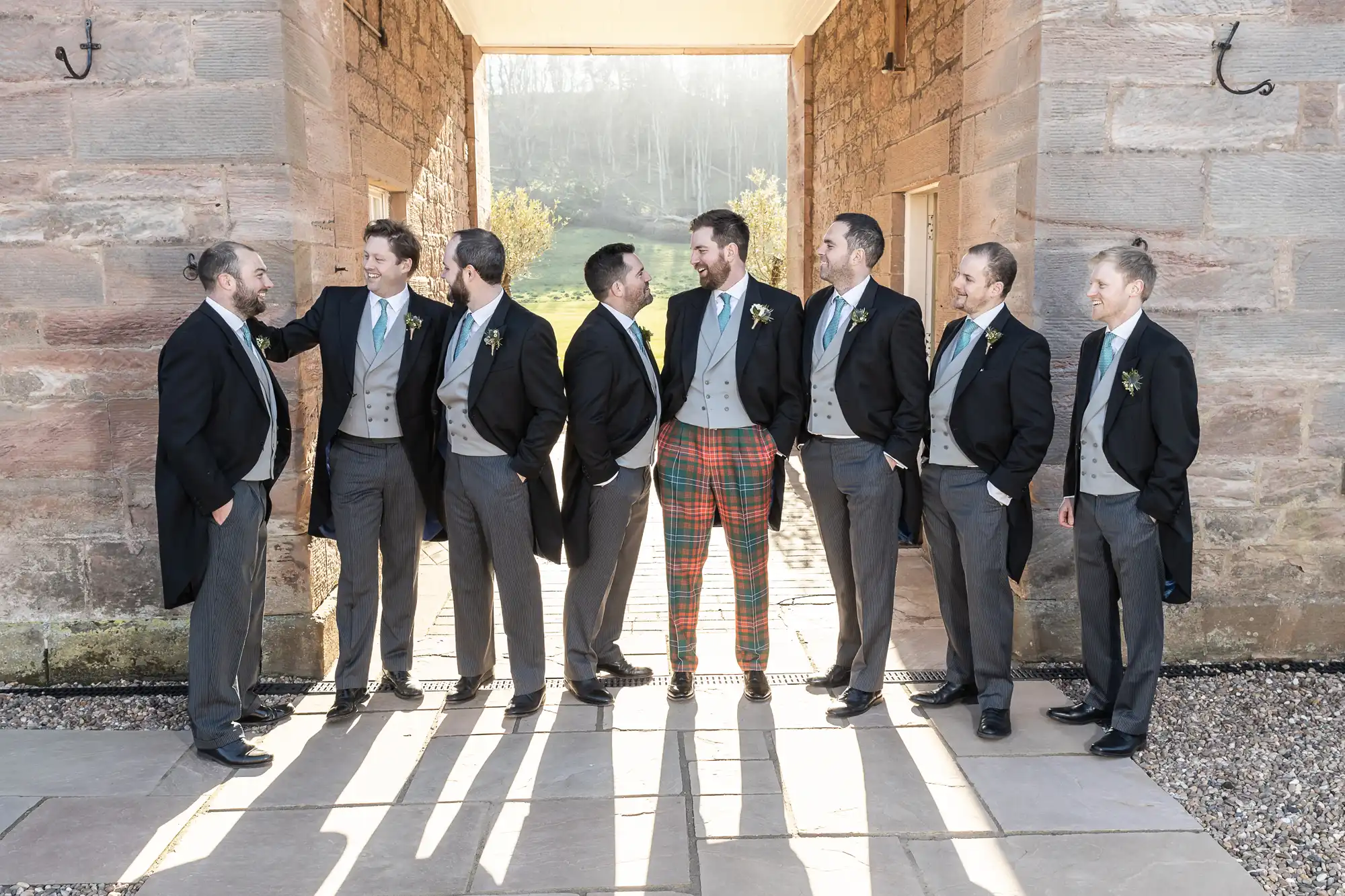 Eight men in formal attire, mostly wearing dark jackets and grey trousers, with one in plaid trousers, standing in a courtyard and smiling at each other.