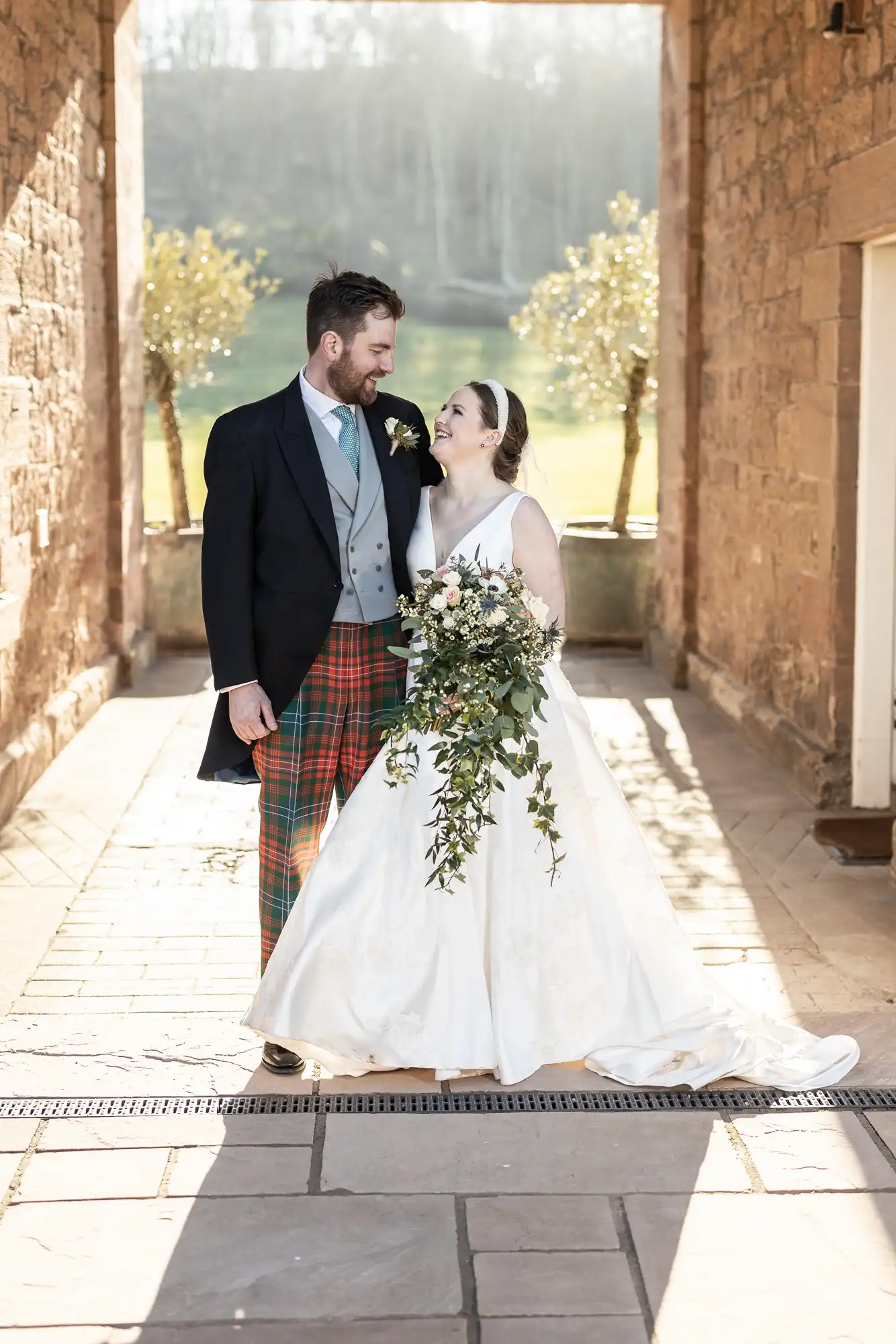 A couple in wedding attire stands in a sunlit brick corridor. The groom wears a suit with tartan trousers, and the bride is in a white gown holding a cascading bouquet. Both are smiling at each other.