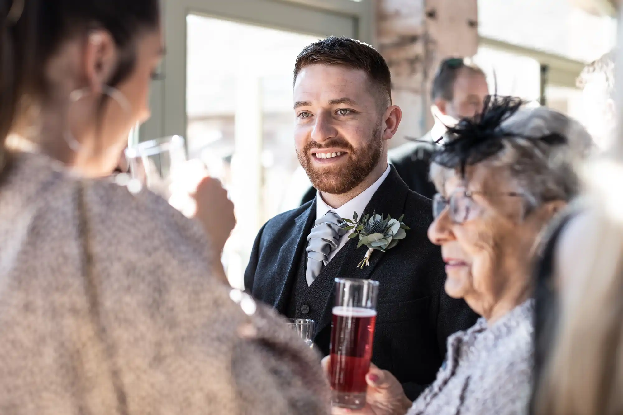 A man in formal attire smiles while holding a drink, standing next to an elderly woman and another person with their back to the camera at a social gathering.