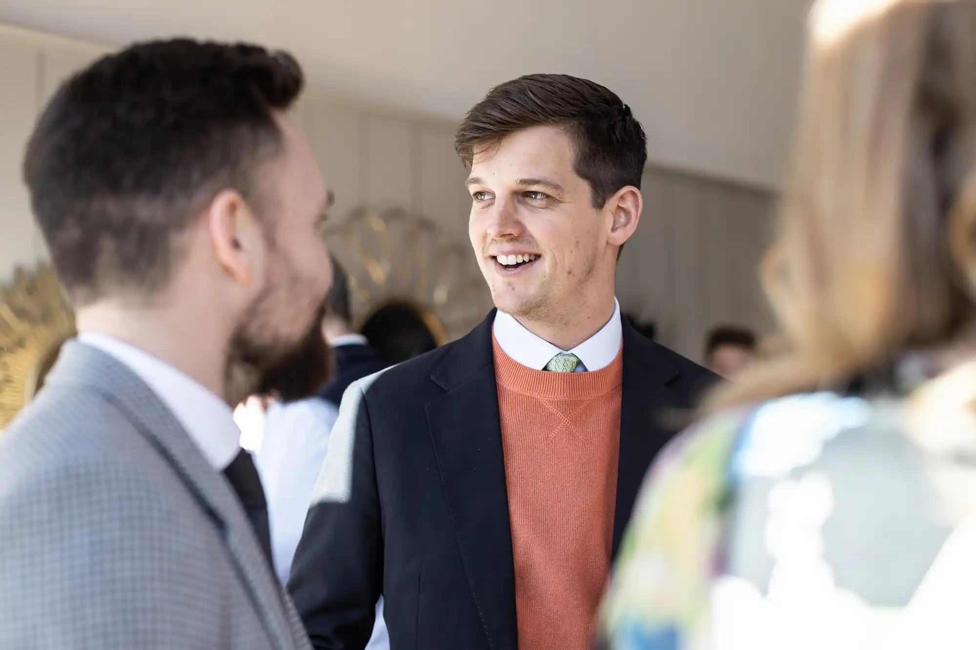 Three people are having a conversation in a well-lit room. One man, centered and wearing a suit with an orange sweater, is smiling. The focus is mainly on him. The others are partially visible.