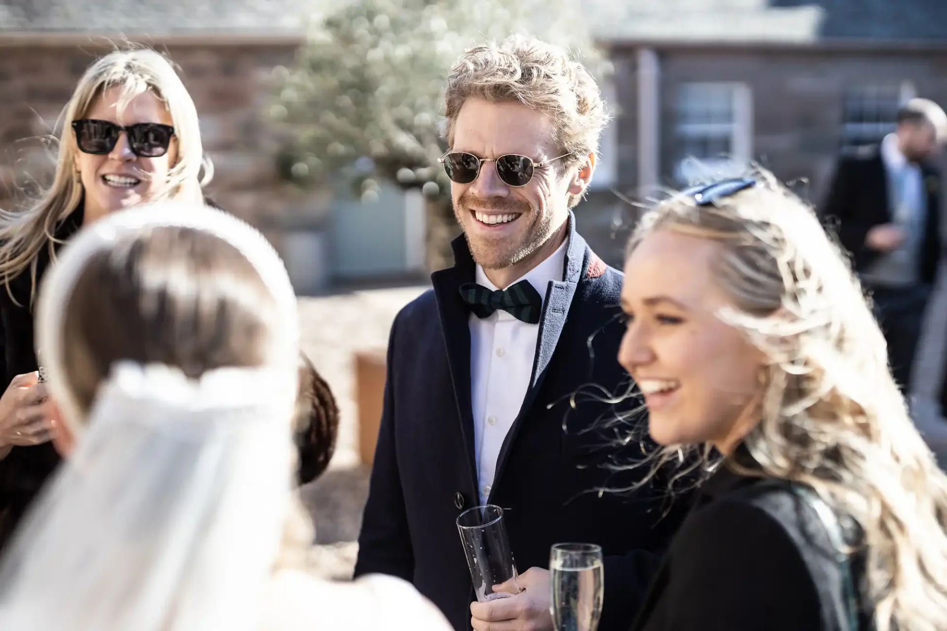 A group of people, dressed formally, engage in conversation outdoors. One man wears sunglasses and a bow tie, holding a champagne flute. In the foreground, a bride with a veil is partially visible.