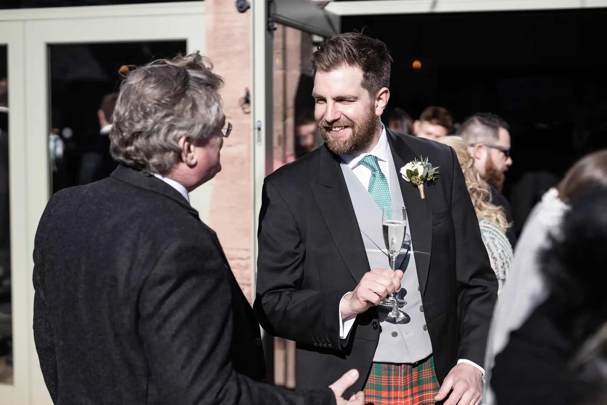 Two men in formal attire engage in conversation, one holding a champagne glass. The setting appears to be an outdoor event.