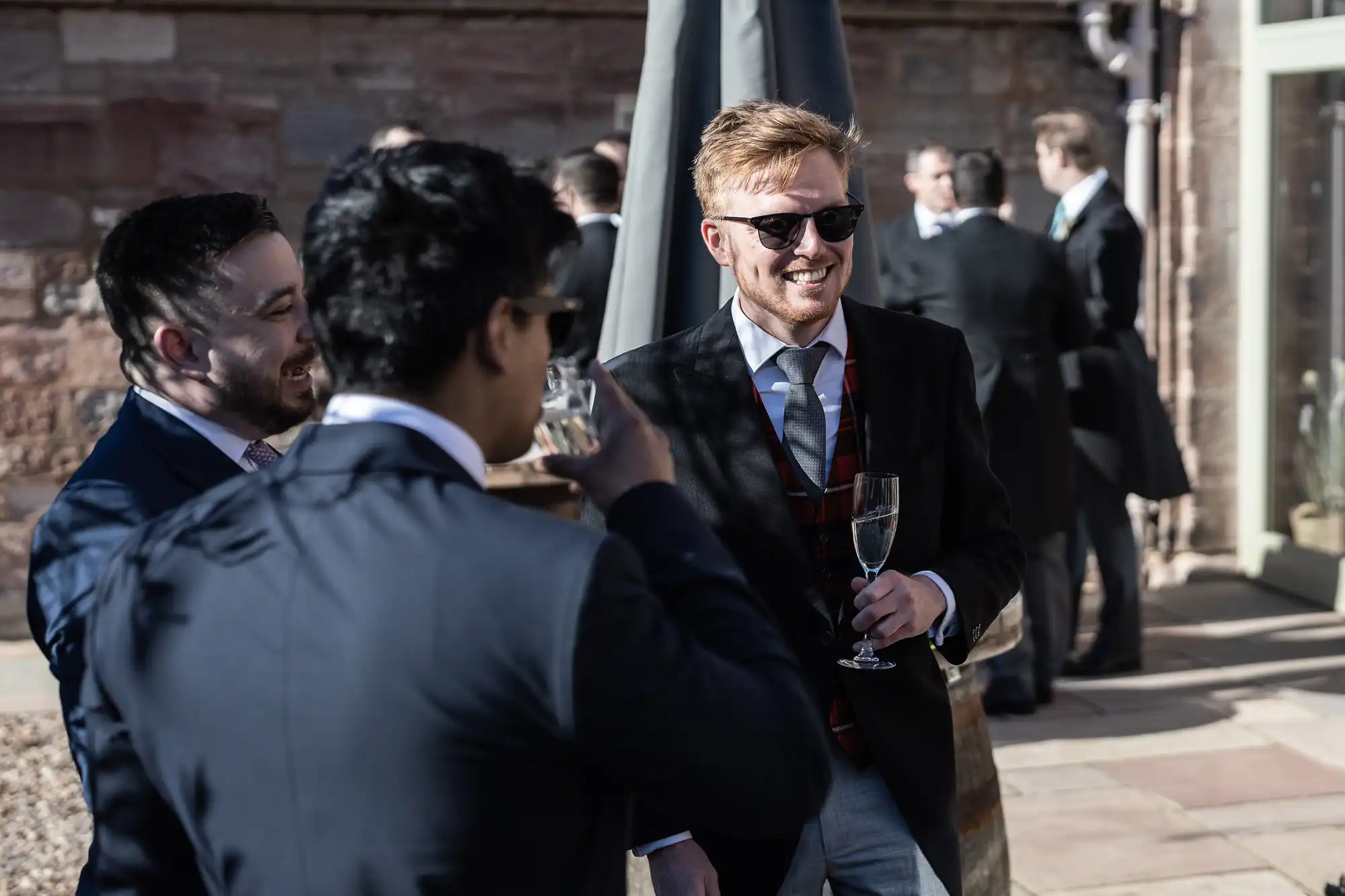 Three men in suits are standing outside, conversing and holding champagne glasses. One man is wearing sunglasses and smiling. Others are in the background.