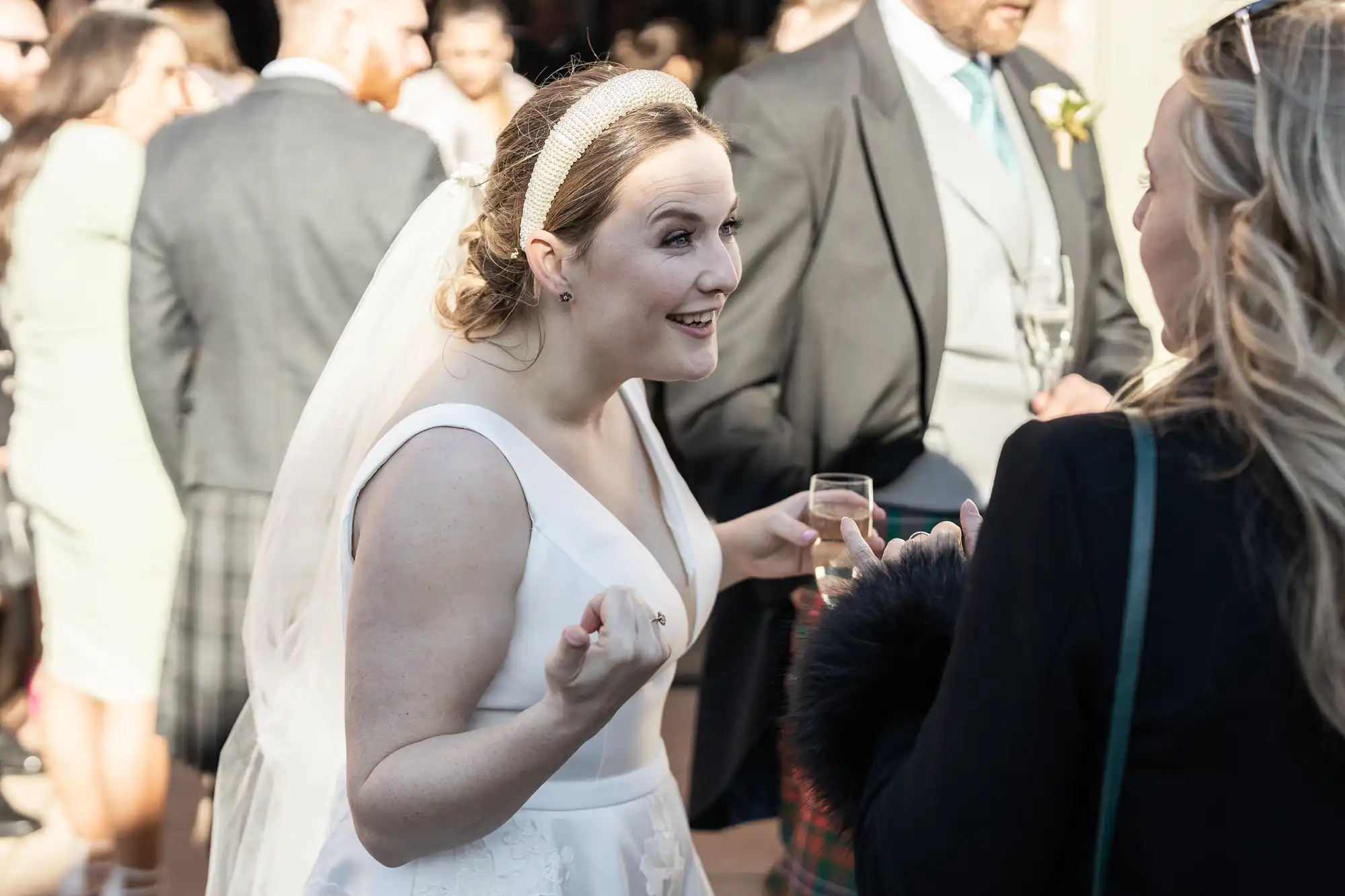 A bride in a white dress and veil, holding a glass, is engaged in a lively conversation with a guest at an outdoor event. Other guests in formal attire are visible in the background.