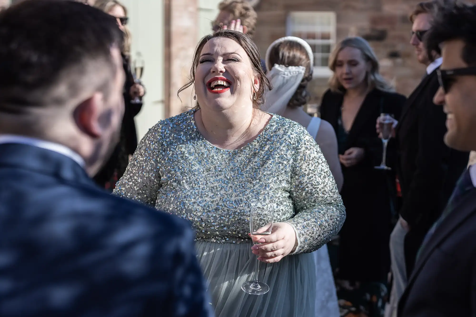 A woman in a sparkling dress laughs and holds a champagne flute at an outdoor gathering. Other people in formal attire are in the background.