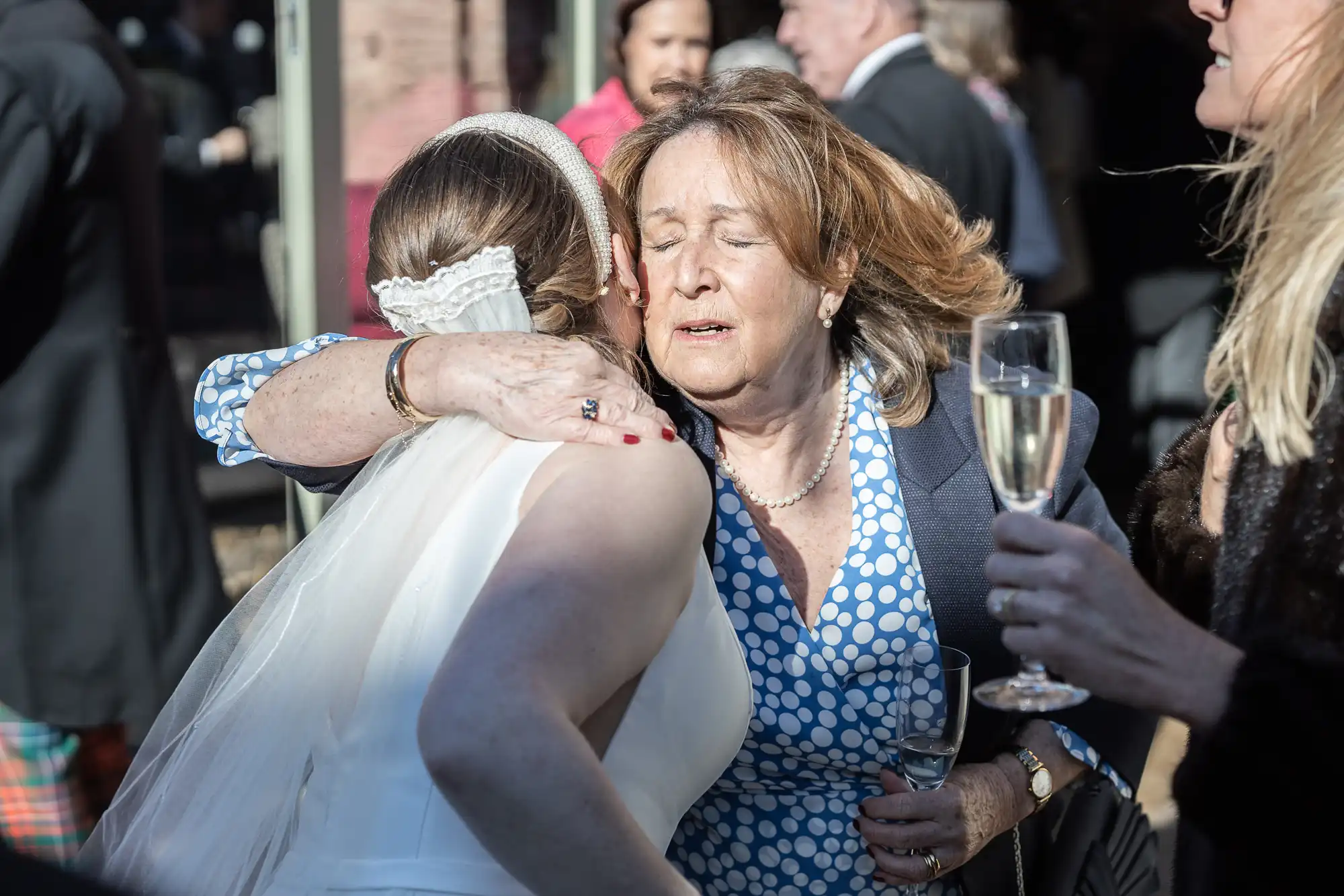 A bride in a white dress embraces an older woman in a blue polka dot dress at an outdoor event. The older woman holds a glass of champagne in one hand.