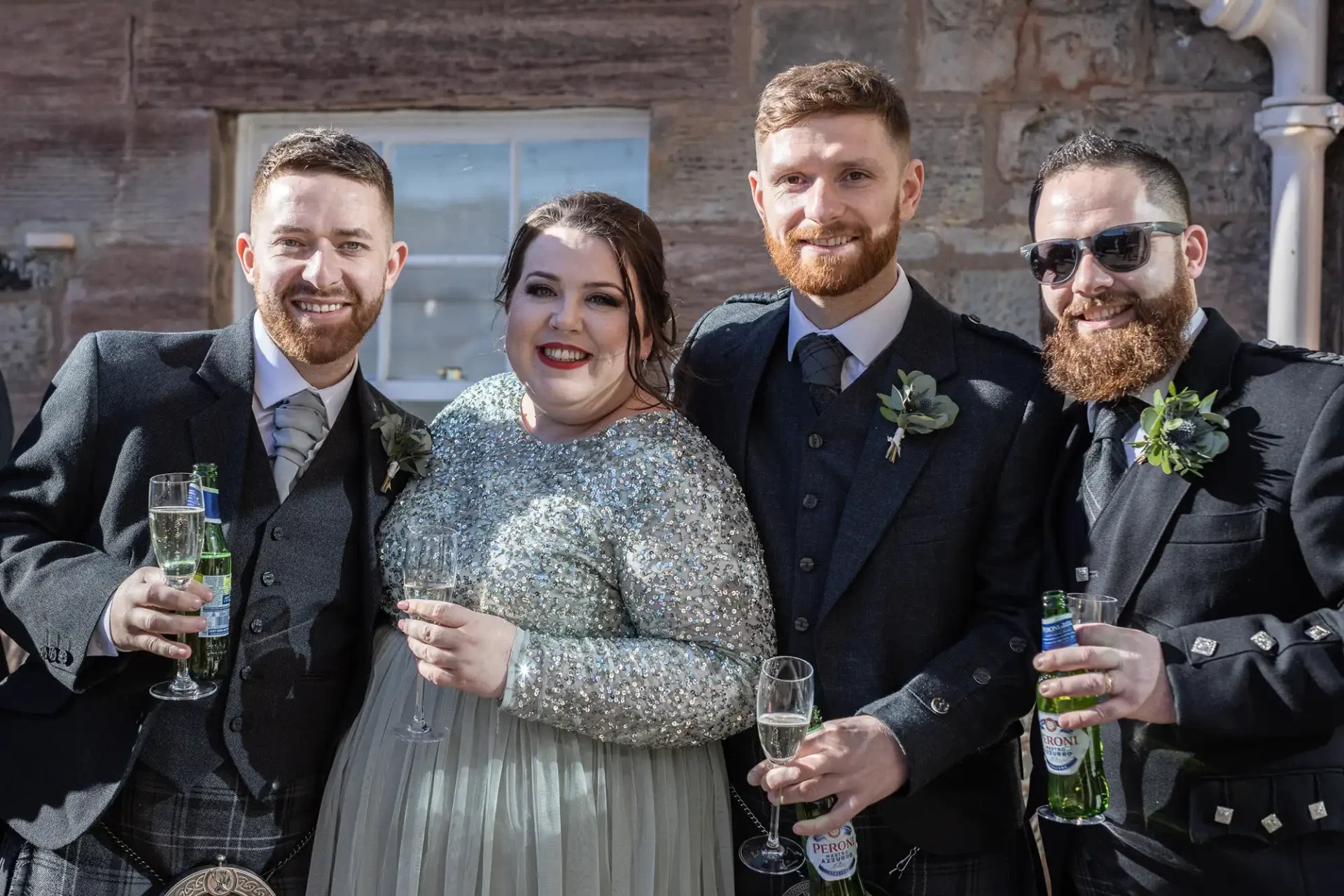 Four people dressed in formal attire standing together, three men and one woman, all holding beverage glasses. The group appears to be at a celebratory event, possibly a wedding, with a brick wall in the background.